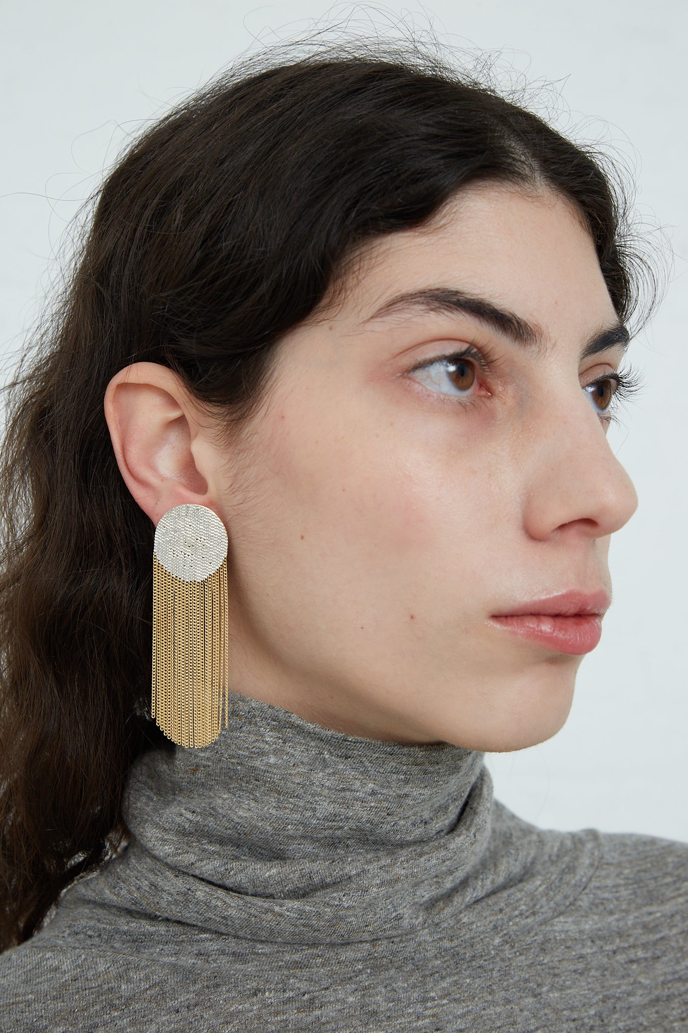 A woman wearing a gray turtleneck and Dot Earrings in Brass Chain and Silver Solder by Hannah Keefe, with brass chain fringe dangling elegantly from her ears.