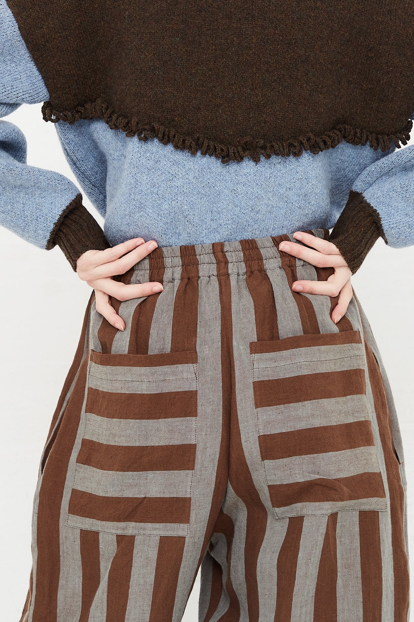 A model wearing a high waist trouser in a striped Irish linen. Brown and blue stripe. Features an elasticated waist and a wide straight leg. Up close and back view of model highlight up close pant and sweater. Showing both pocket placements and details. Pockets feature horizontal stripe in contrast to vertical pant stripe. Designed by Cawley - Oroboro Store