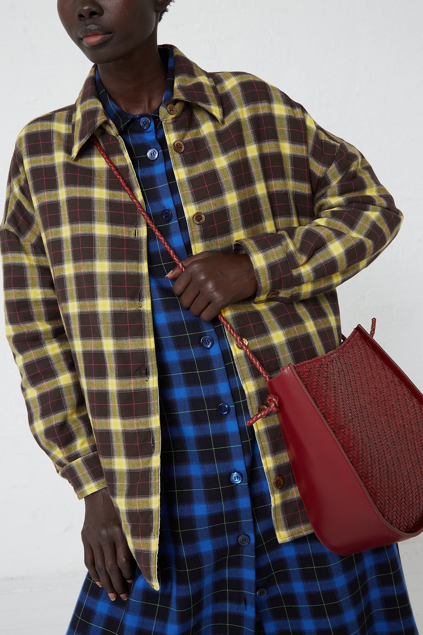 A woman wearing a plaid shirt and carrying a Dragon Diffusion Wanaka Crossbody Bag in Bordo made of buffalo leather using handwoven techniques.