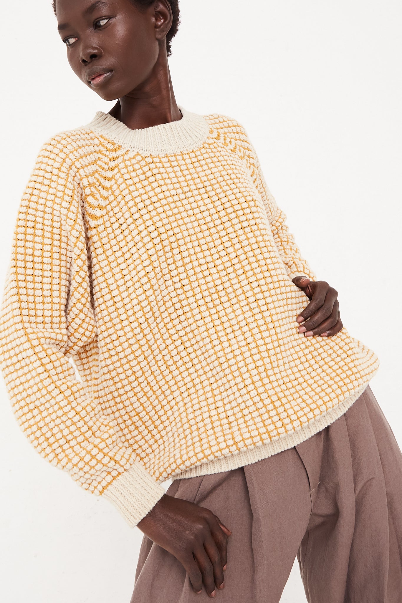 The model is wearing an oversized Lalin Crewneck Sweater in Natural Gold checkered made of merino wool by Jan-Jan Van Essche.