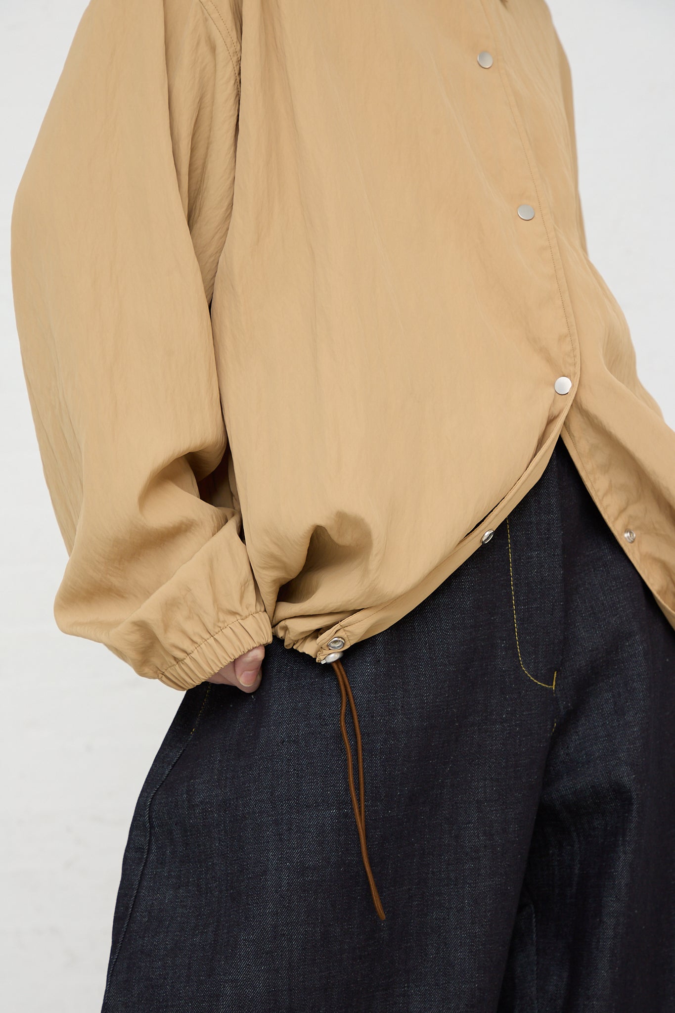 A woman wearing a tan Studio Nicholson Sprung Coach Jacket in Sand and jeans.