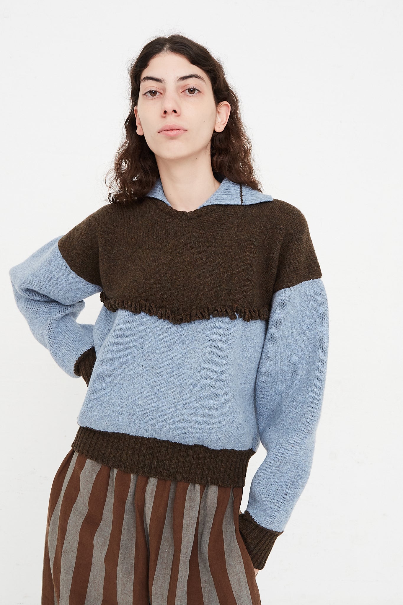 A model wearing a knit pullover sweater in a British wool. Features a polo collar with dropped shoulders and contrast color on top, cuffs and hem. Front view and up close. Showcasing sleeves, details and pants. One hand in pocket. Designed by Cawley - Oroboro Store