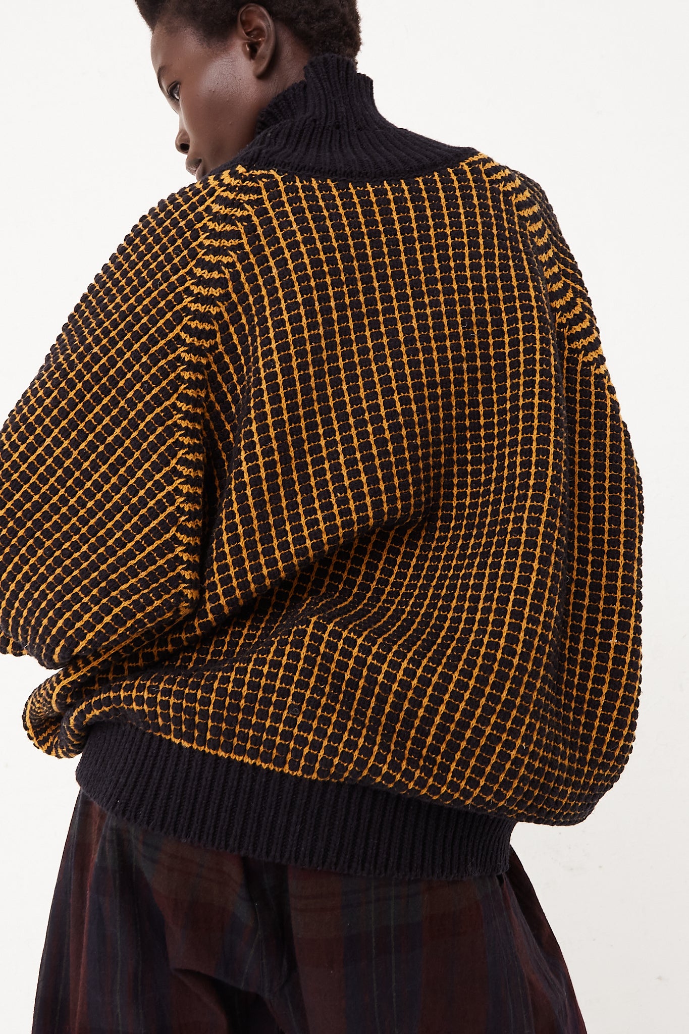 The back view of a woman wearing a yellow and black checkered Turtleneck Sweater in Pitch Black Gold Lalin made from merino wool by Jan-Jan Van Essche.