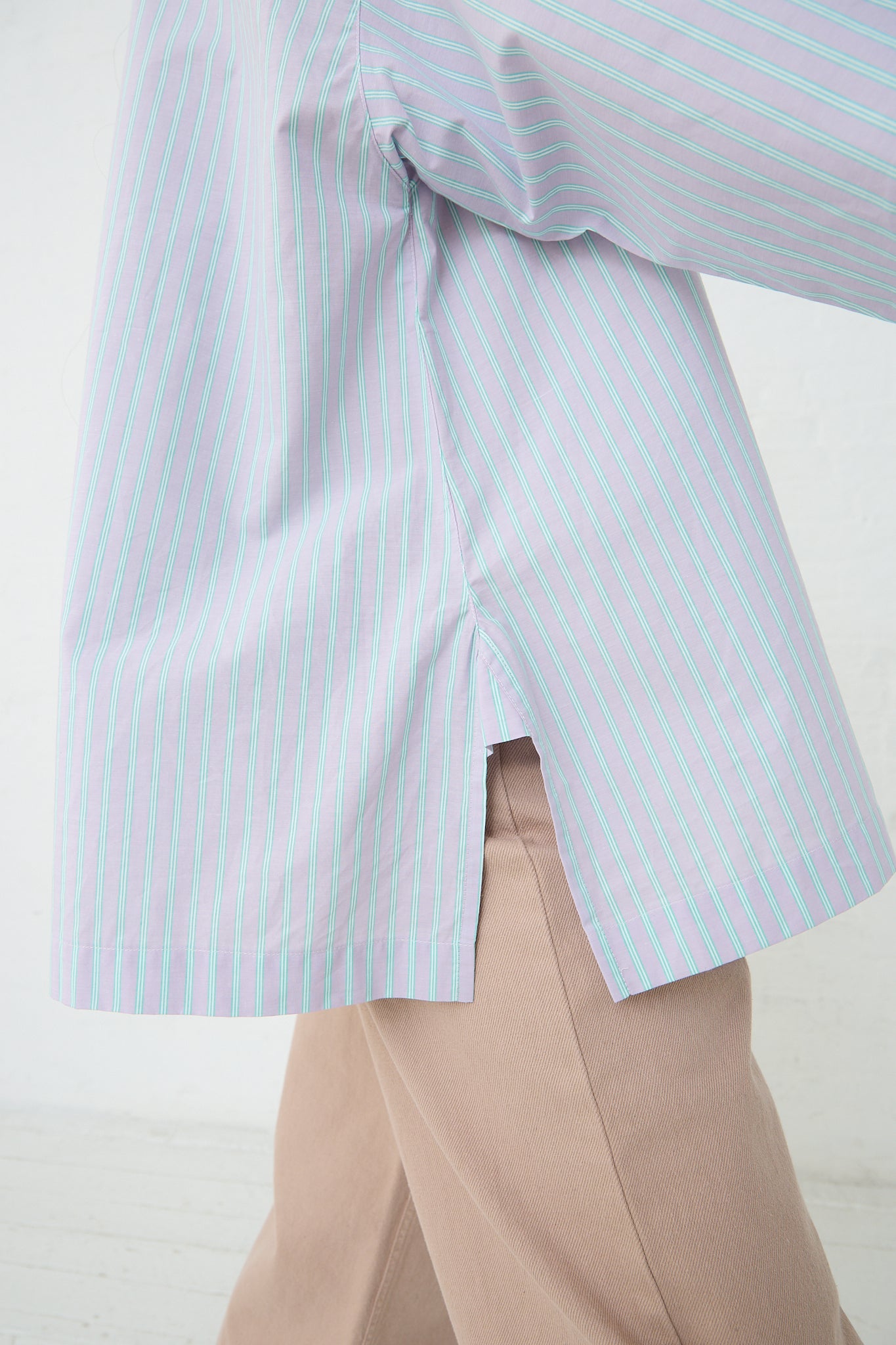 A woman wearing a Baserange Giza Cotton Kolla Shirt in Lilac Stripe with pink pants. Side view to highlight bottom of shirt.