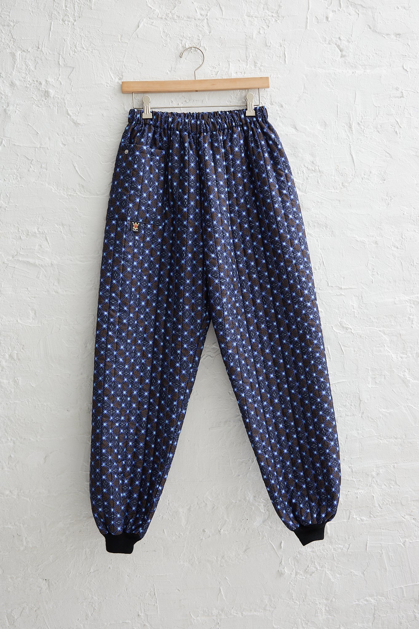 A pair of Monpe Pant No. 08 in Floral Print B - S by Bless, blue floral printed nylon jogging pants with an elasticated waist, hanging on a white wall.