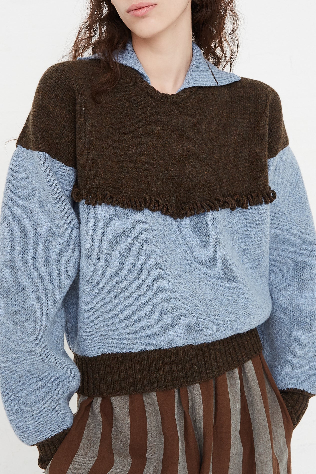 A model wearing a knit pullover sweater in a British wool. Features a polo collar with dropped shoulders and contrast color on top, cuffs and hem. Front view and up close. Showcasing details and pants. Hands in pockets. Designed by Cawley - Oroboro Store