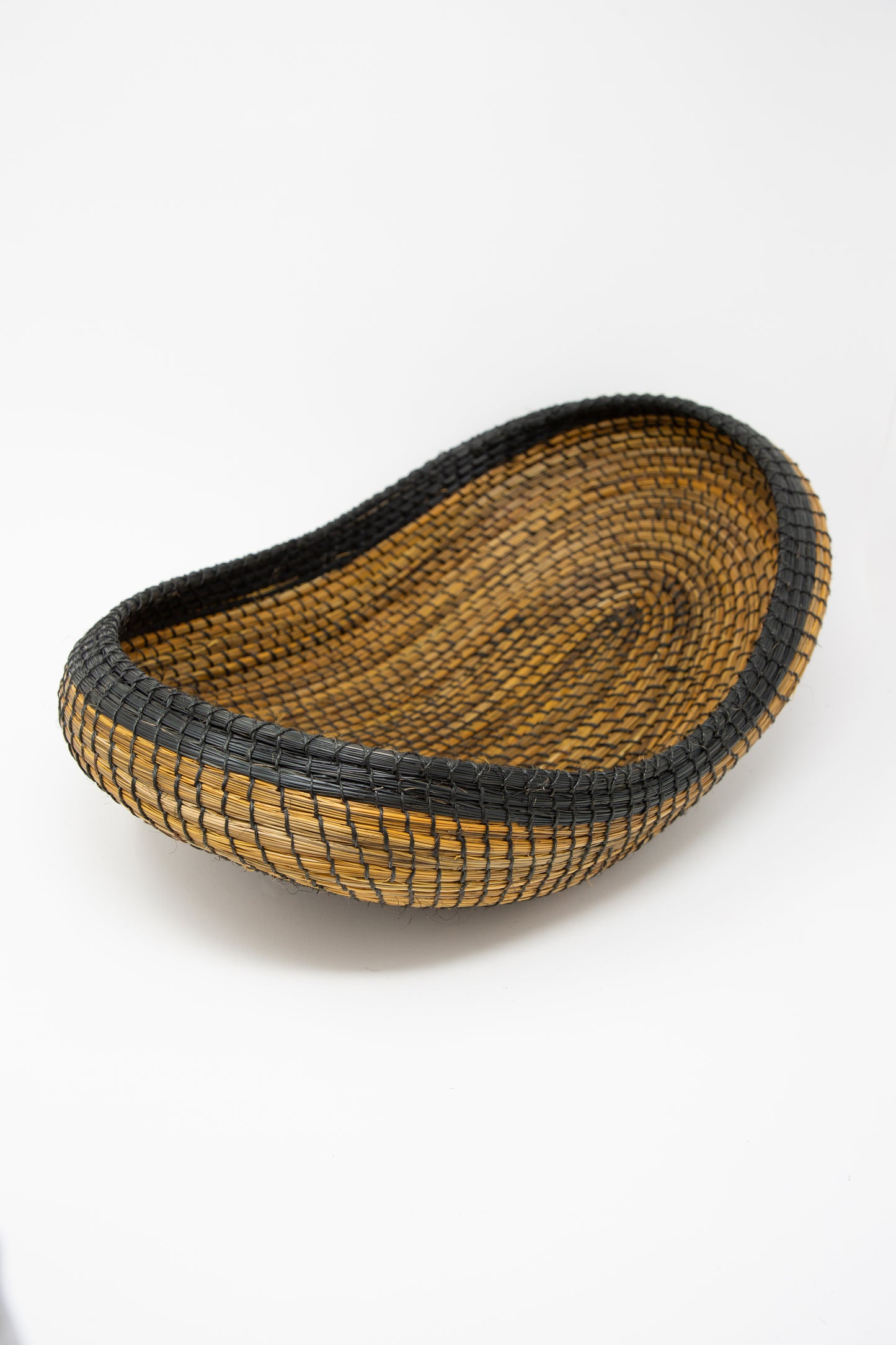 A black and brown Asopafit Canoe Basket by Plaza Bolivar, showcasing traditional weaving skills from Colombia, sits on a white surface.