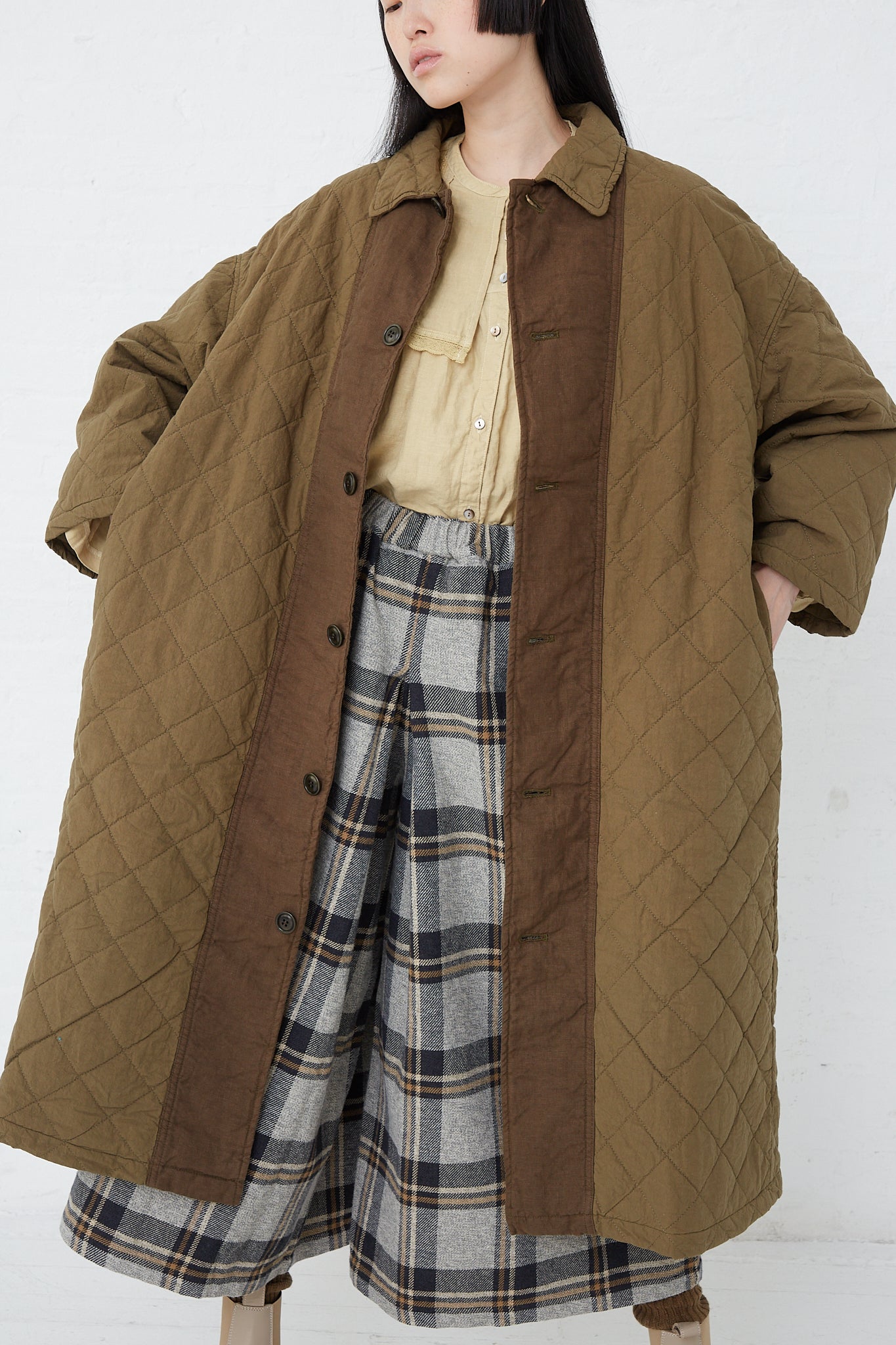 A woman wearing an oversized nest Robe Cotton Ramie Quilted Long Coat in Olive and plaid skirt.