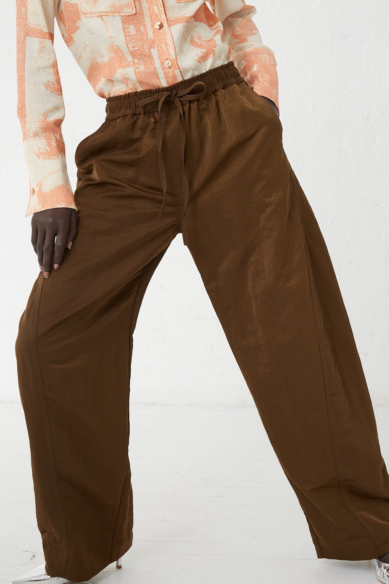A model in Rejina Pyo's Nylon Una Trousers in Brown with contrast stitching and an elasticated waist.
