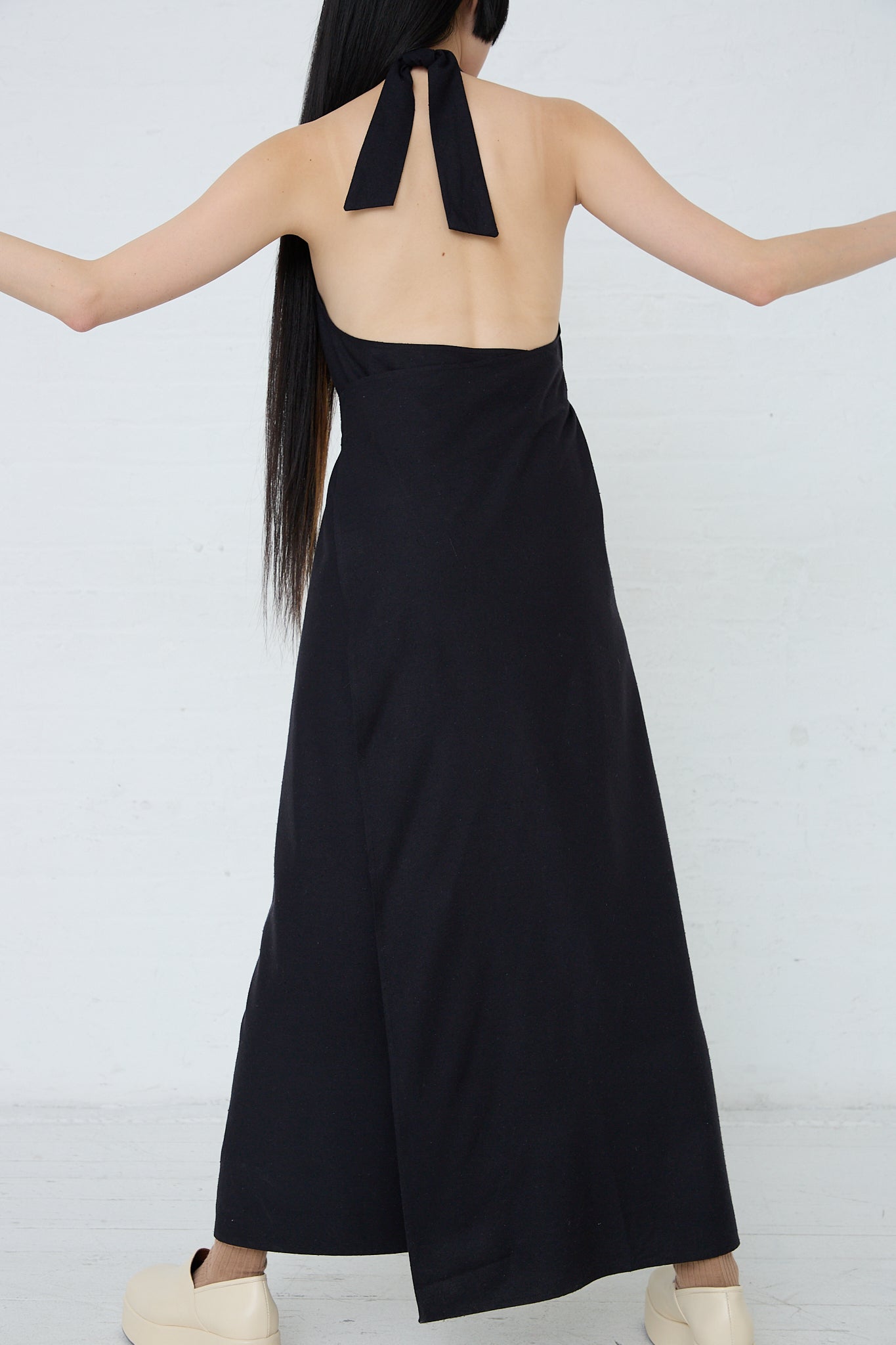 The back view of a woman in a Wild Silk Trope Apron Dress in Black with her arms outstretched.
