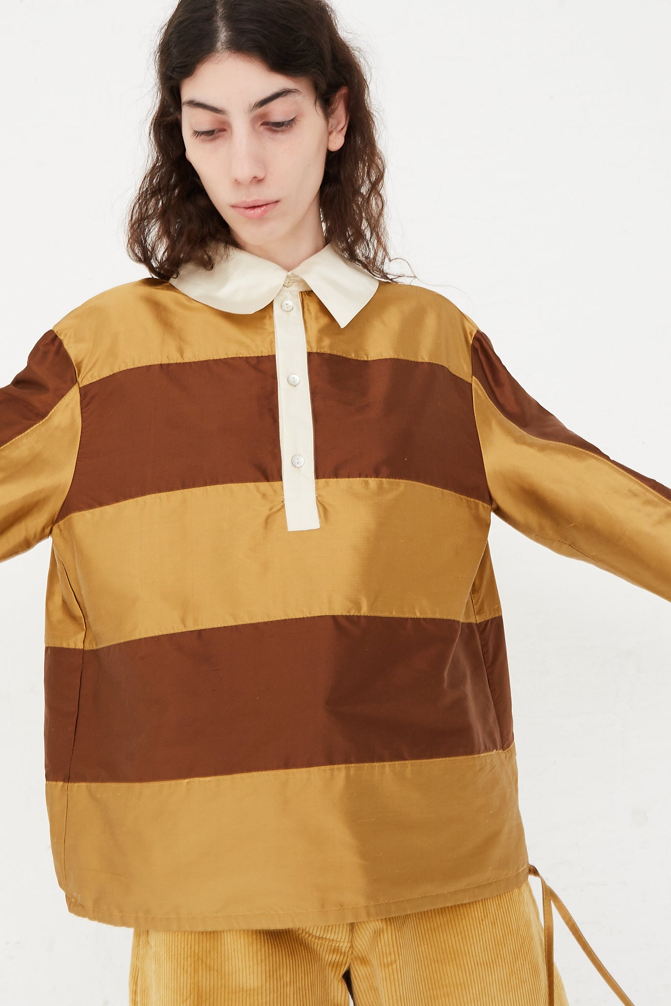 A model wearing a striped rugby shirt in a shiny silk doupion. Features a contrast fold-over collar and button placket, with an adjustable drawstring at hem. Front view and up close. Designed by Cawley - Oroboro Store 