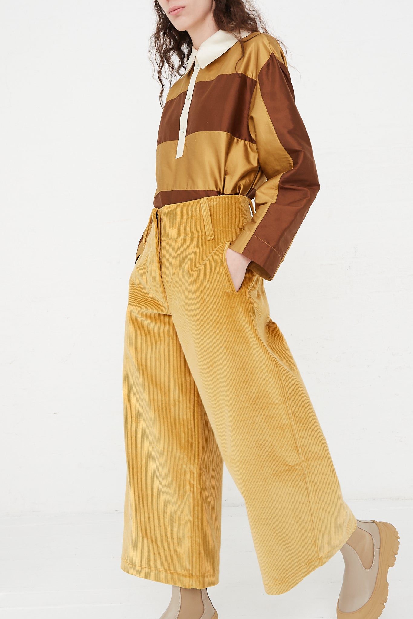 A model wearing a high waist trouser in a British cotton cord. Features a wide waistband, pleat detail and side welt pockets. Side view and full length. Designed by Cawley - Oroboro Store