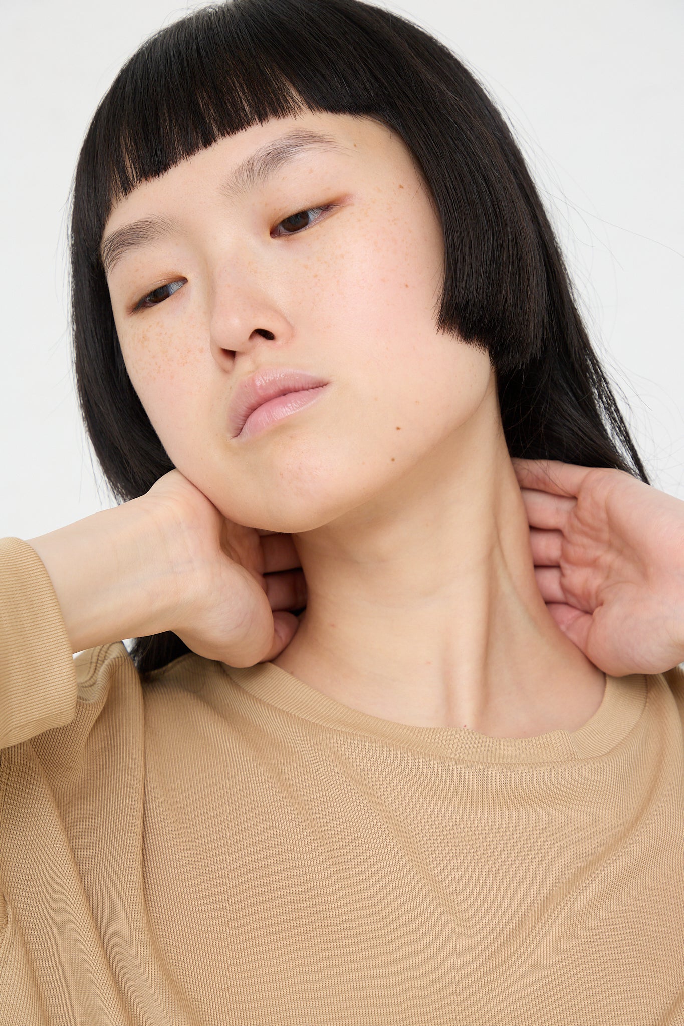 A woman wearing a Studio Nicholson Simmons Long Sleeve Top in Sand with dropped shoulders and black hair.
