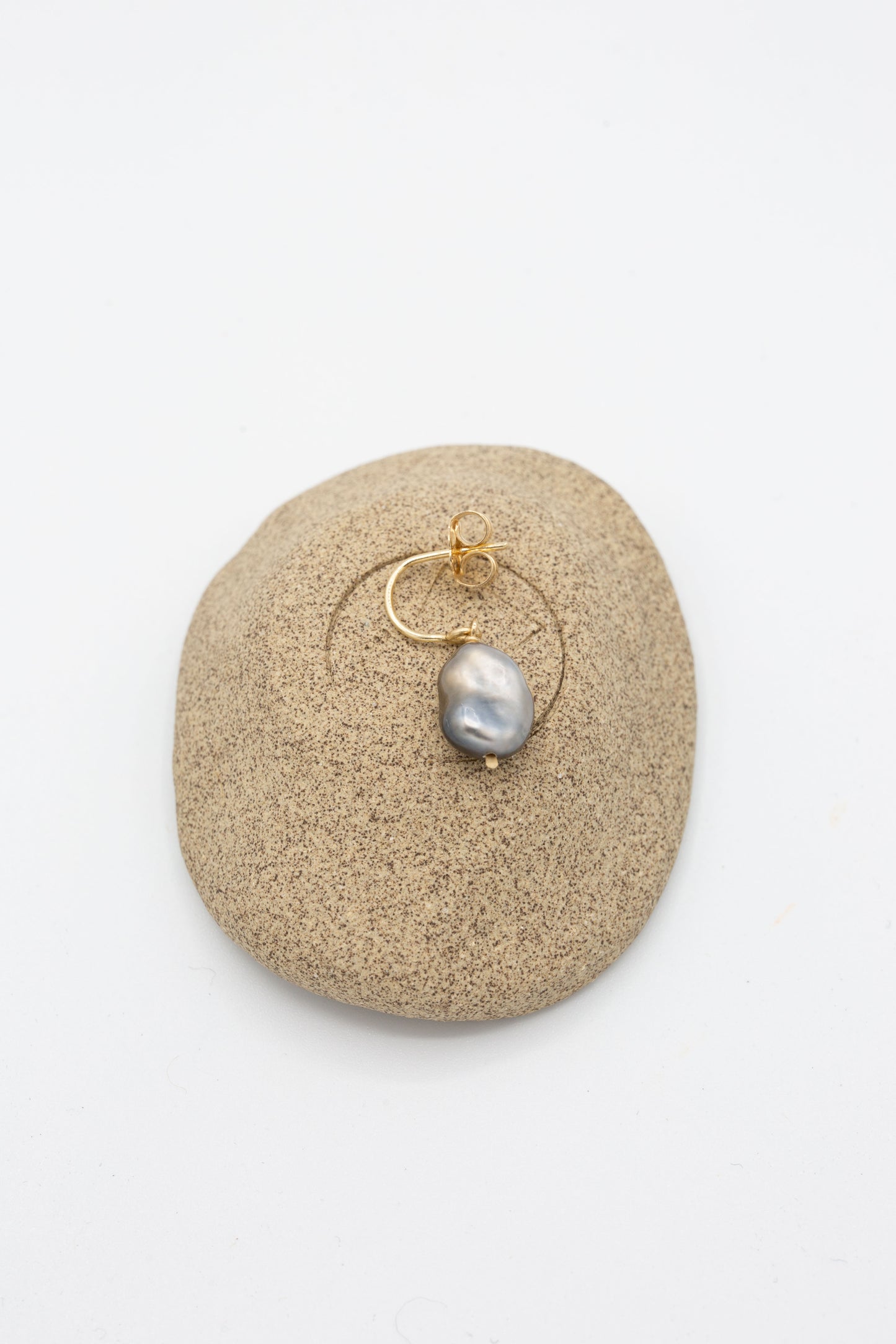 A Mary MacGill 14K Crescent Charm Earrings in Grey Keshi Pearl adorned with a Keshi pearl. Laying on a beige colored stone.