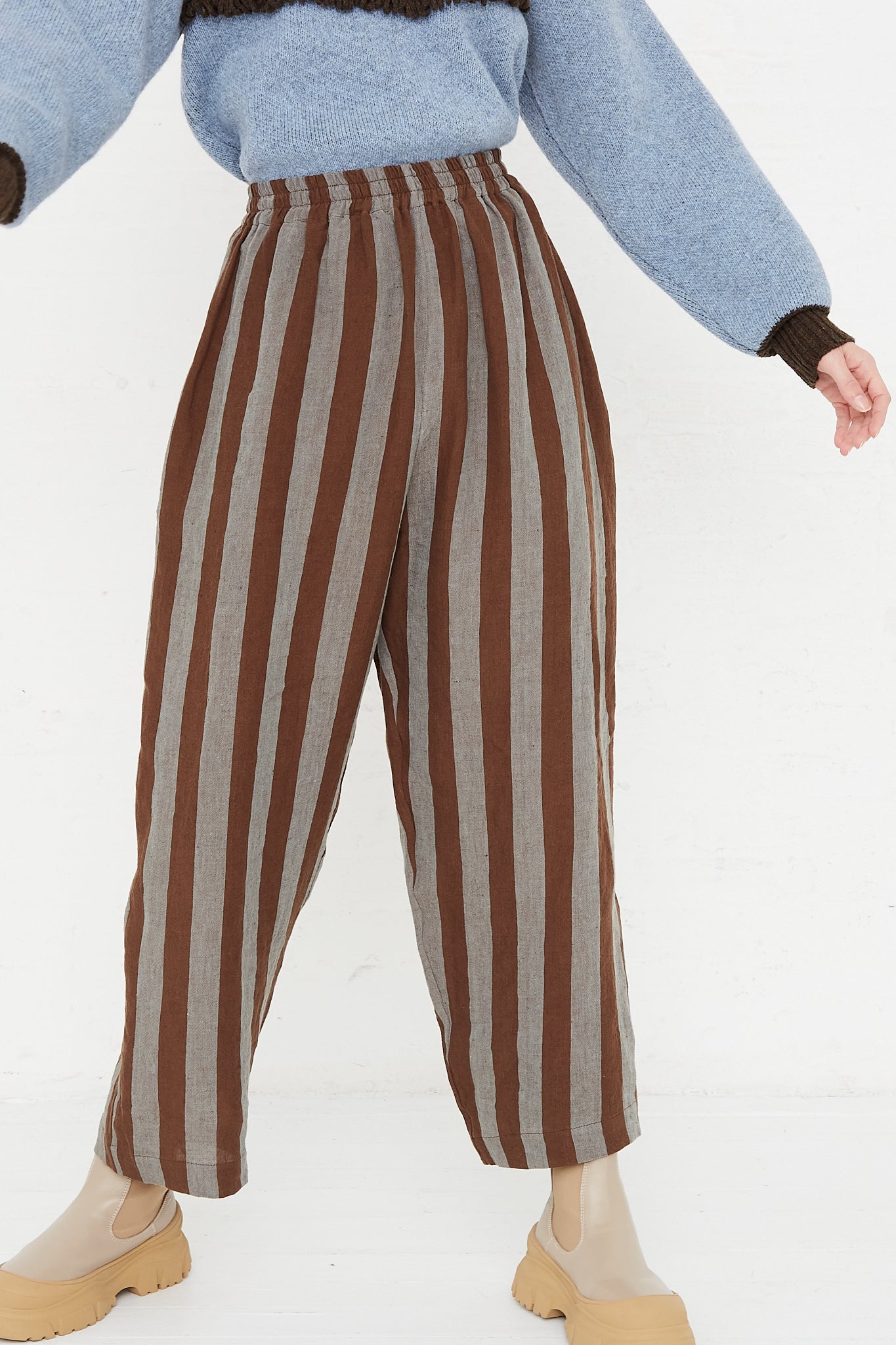 A model wearing a high waist trouser in a striped Irish linen. Brown and blue stripe. Features an elasticated waist and a wide straight leg. Front and upclose view of model highlight details of pant, bottom and sleeves of sweater and boots. Designed by Cawley - Oroboro Store