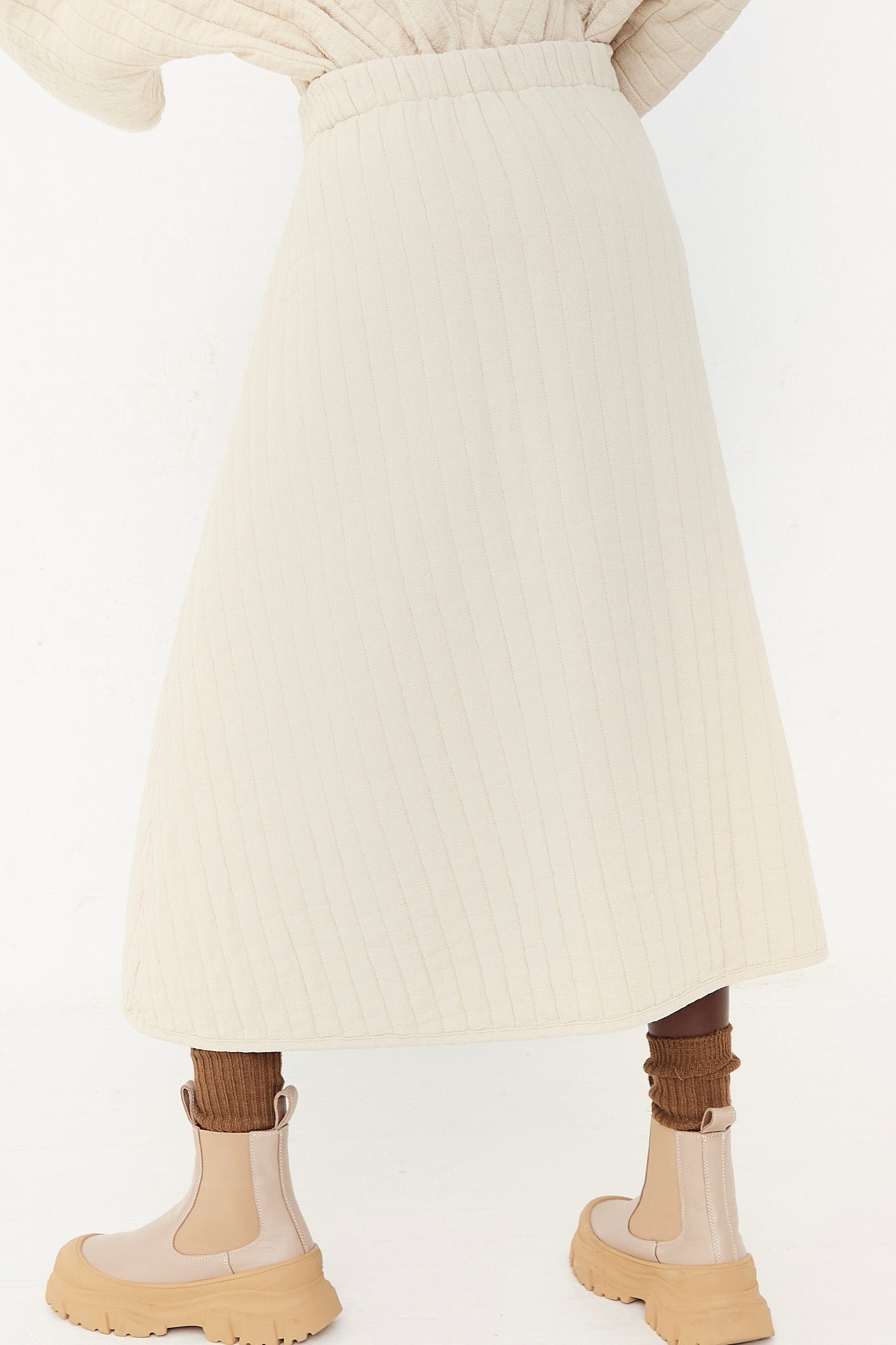 The back of a woman wearing a cream Black Crane sweater and tan boots, with an elasticated waist, in Ivory Quilted Skirt.