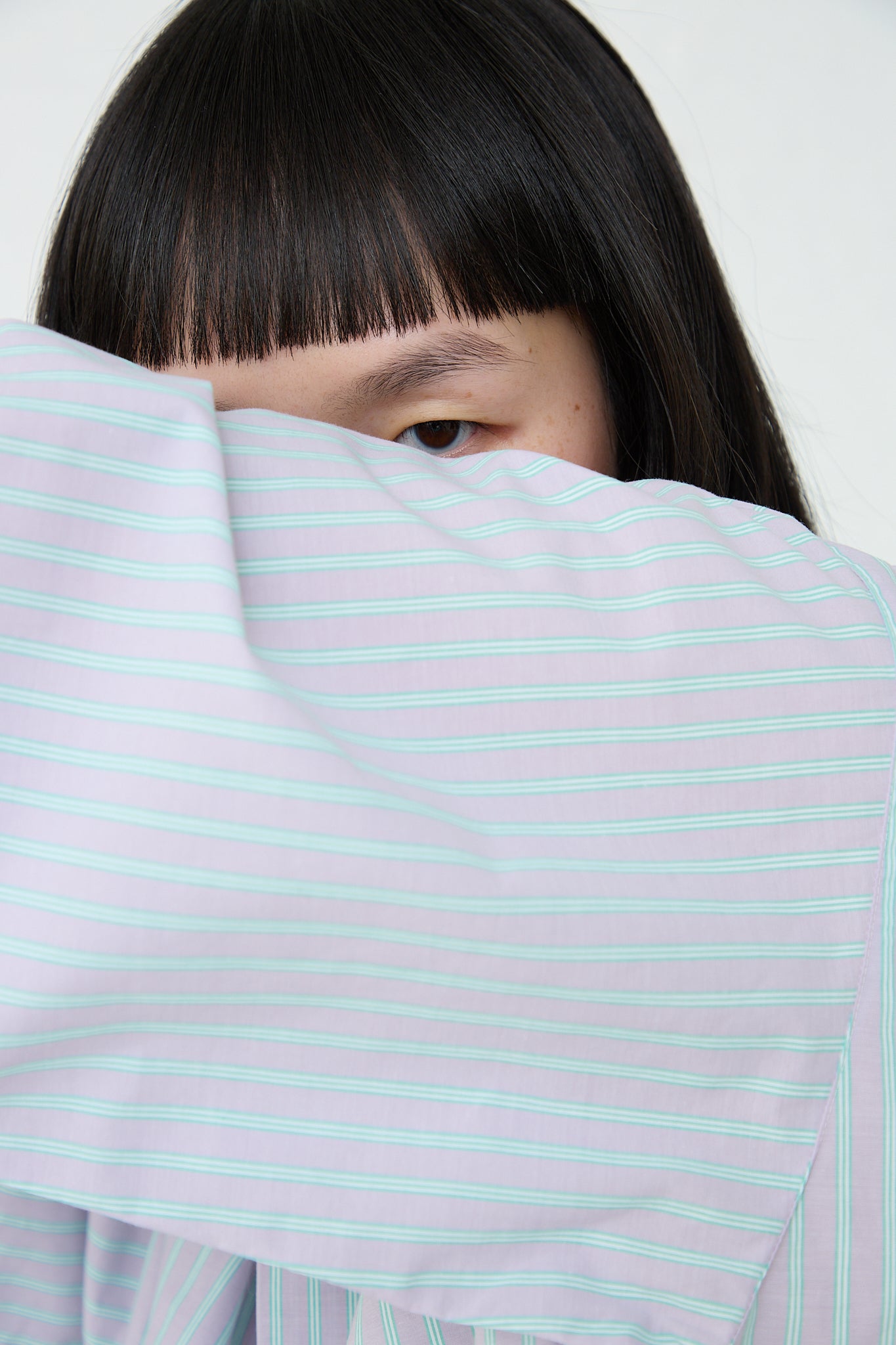 A woman wearing a Giza Cotton Kolla Shirt in Lilac Stripe by Baserange covering her face to highlight stripe details.