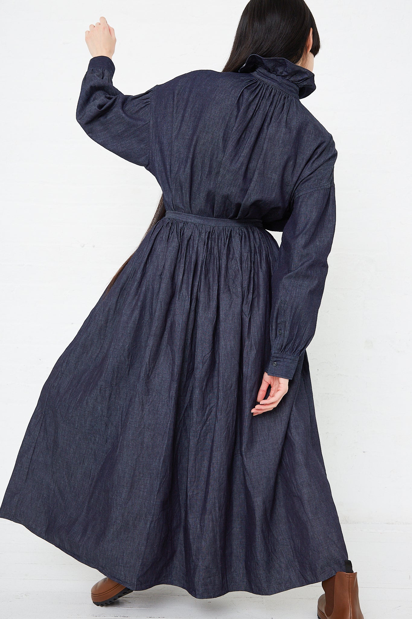 The back of a woman wearing a long Toujours Cotton Denim Cloth Pleated Maxi Skirt in Indigo, made from organic cotton denim, featuring side seam pockets and an A-line skirt.