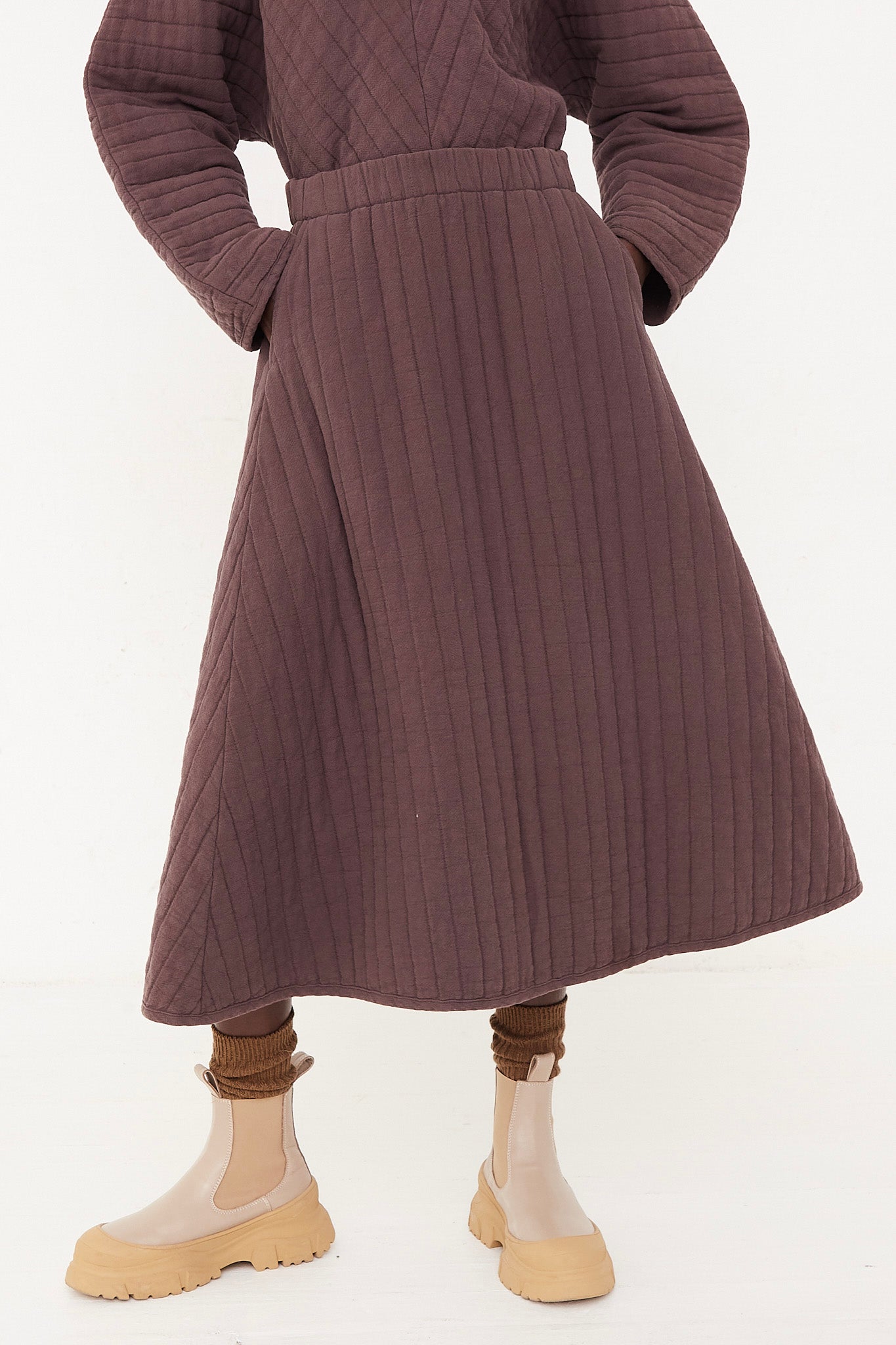 The model is wearing a Black Crane Quilted Skirt in Plum with an elasticated waist.
