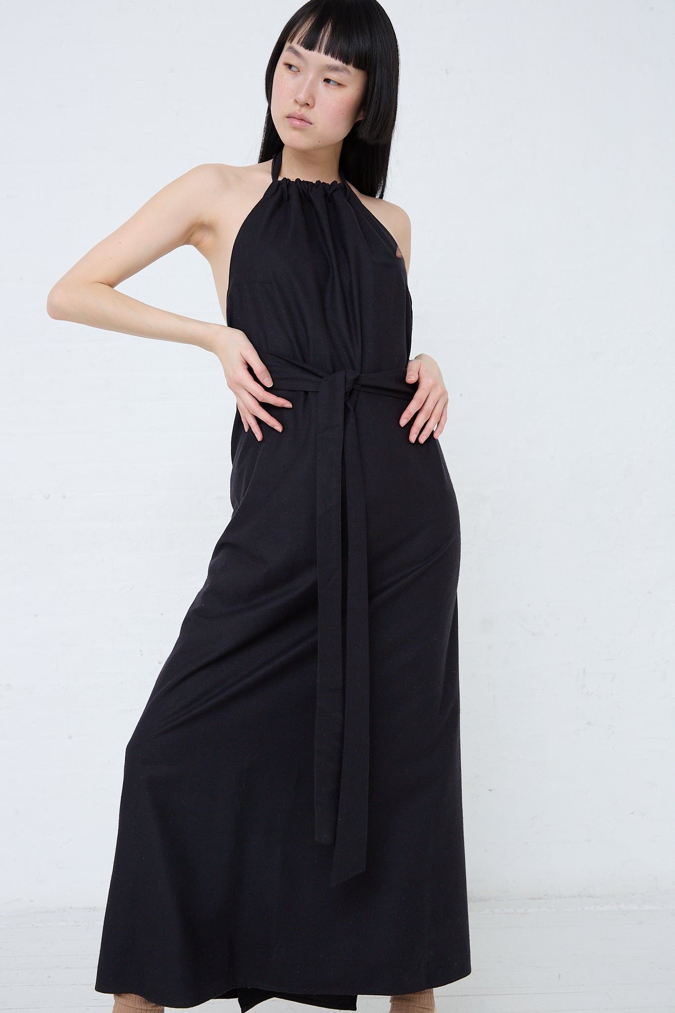 A woman wearing a Wild Silk Trope Apron Dress in Black made by Baserange. Full length and front view.