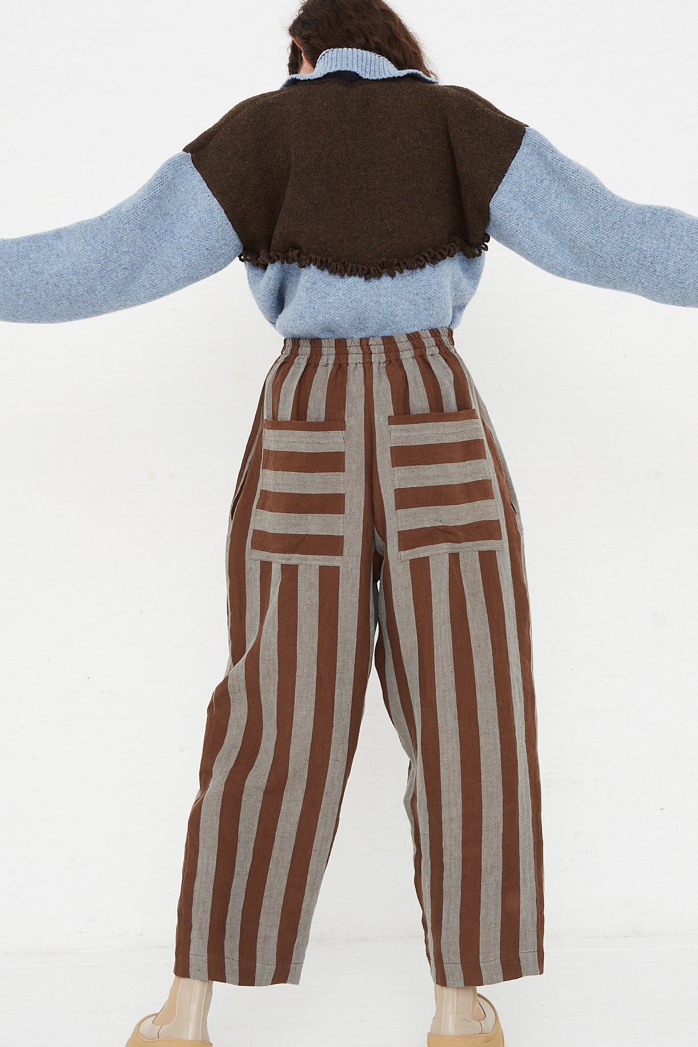 A model wearing a high waist trouser in a striped Irish linen. Brown and blue stripe. Features an elasticated waist and a wide straight leg. Full length and back view of model highlight full pant, sweater and boots. Showing both pocket placements and details. Pockets feature horizontal stripe in contrast to vertical pant stripe. Designed by Cawley - Oroboro Store