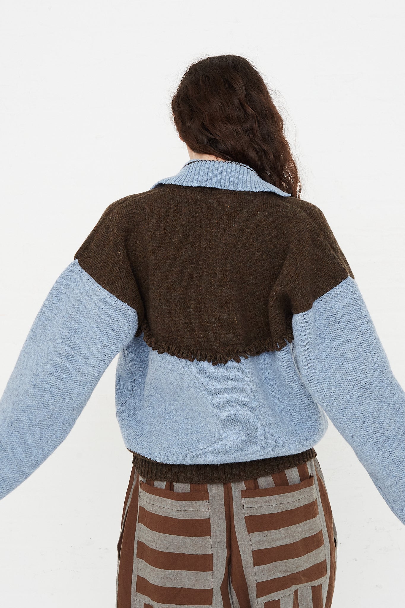 A model wearing a knit pullover sweater in a British wool. Features a polo collar with dropped shoulders and contrast color on top, cuffs and hem. Back view and up close. Showcasing sleeves, back details and pant pockets. Designed by Cawley - Oroboro Store