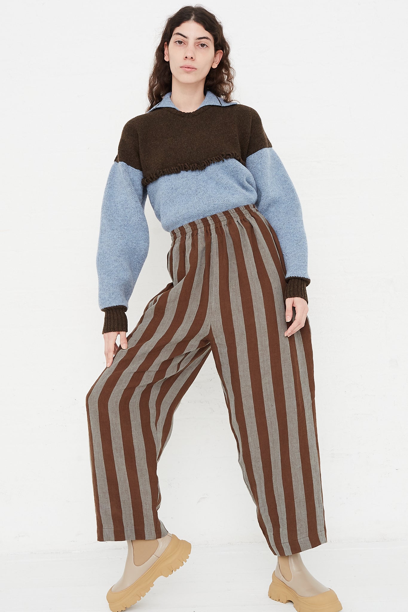 A model wearing a high waist trouser in a striped Irish linen. Brown and blue stripe. Features an elasticated waist and a wide straight leg. Front and full view of model highlight full pant, sweater and boots. Model slightly leaning to show movement of pant. Designed by Cawley - Oroboro Store