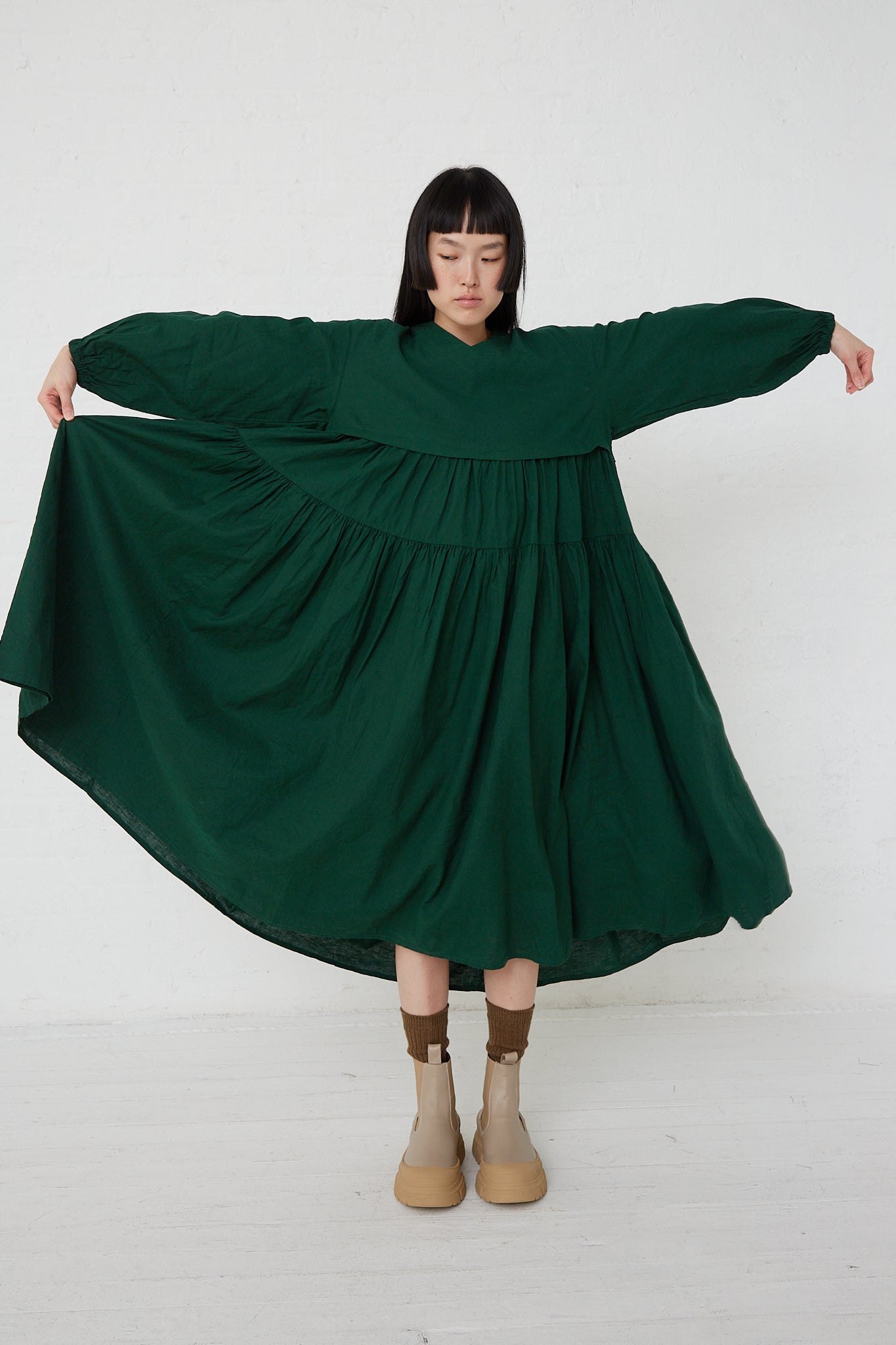 A woman in a UpcycleLino Linen Tiered Gather Dress in Green by nest Robe, made from upcycled linen and cotton blend fabric, is posing elegantly in front of a white wall.