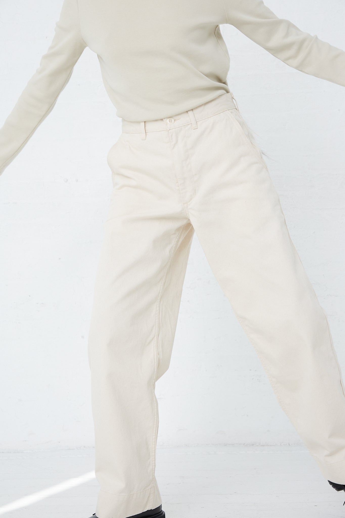 A woman wearing As Ever's 51 Chino in Natural pants and a sweater with corded pocket detail in a white room.