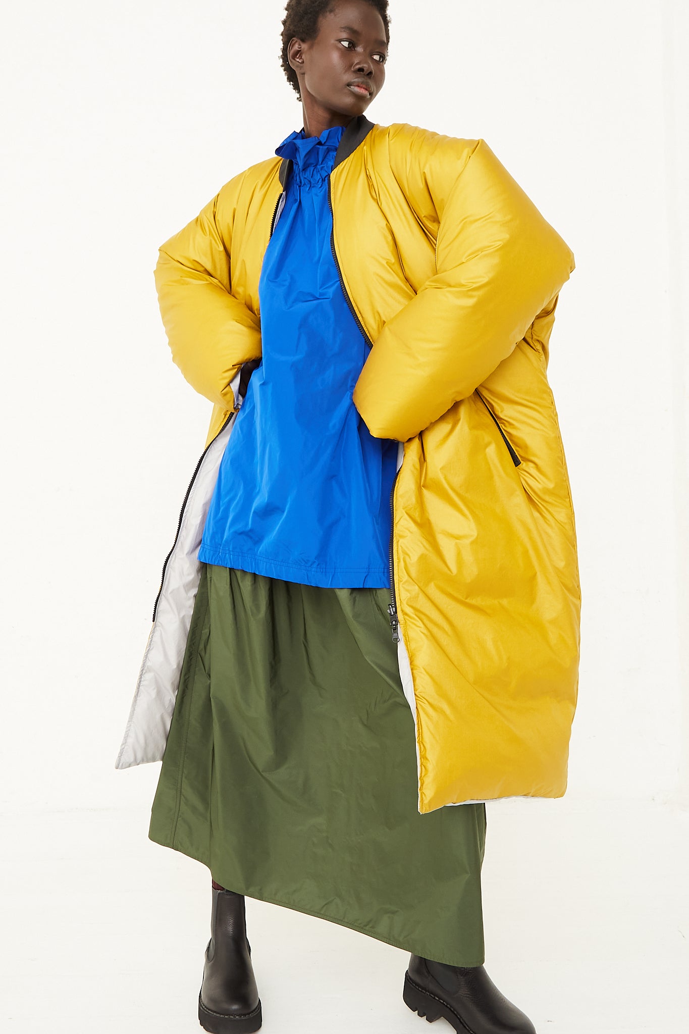 A model wearing an oversized down jacket in a water repellent nylon. Features a bomber collar, zip closure, adjustable ties at hem and a reversible fit. Full length. Coat opened highlighting the two-toned details. Hands on hips.Designed by Sofie D'Hoore - Oroboro Store