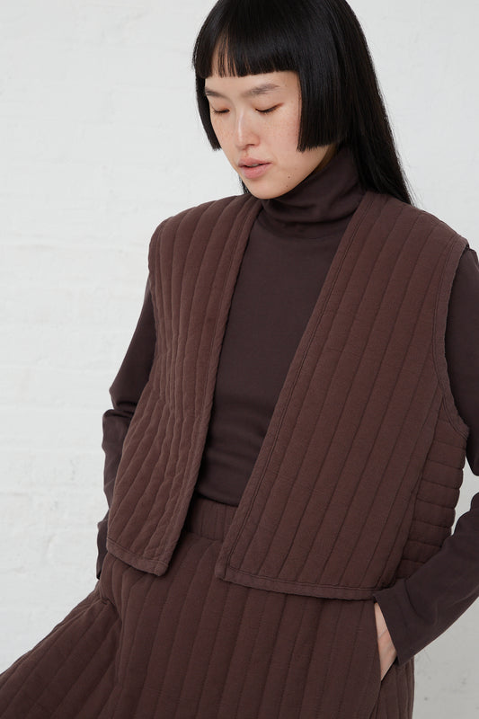 A Black Crane Quilted Vest in Plum presents a woman wearing an open vest; the vest is brown and made of quilted woven cotton.