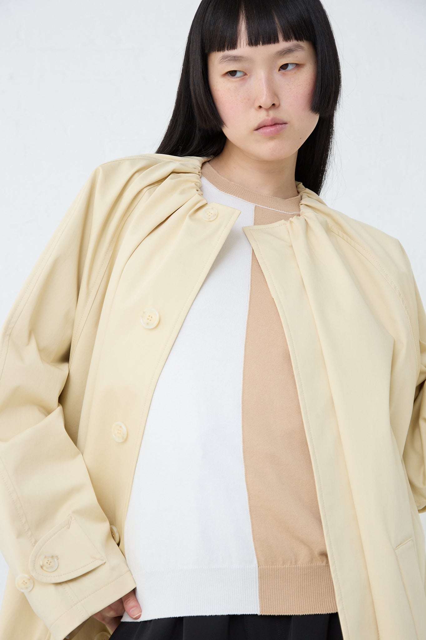 A model wearing a MM6 pale yellow collarless Trench Coat and black pants. Front view. 