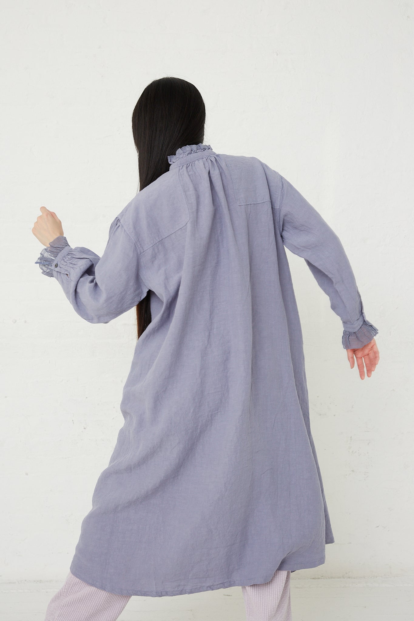 A woman in a blue shirt and pants with a front button closure, is standing in front of a white wall wearing the nest Robe Linen Cotton Lace Omi-Zarashi Dress in Lavender.