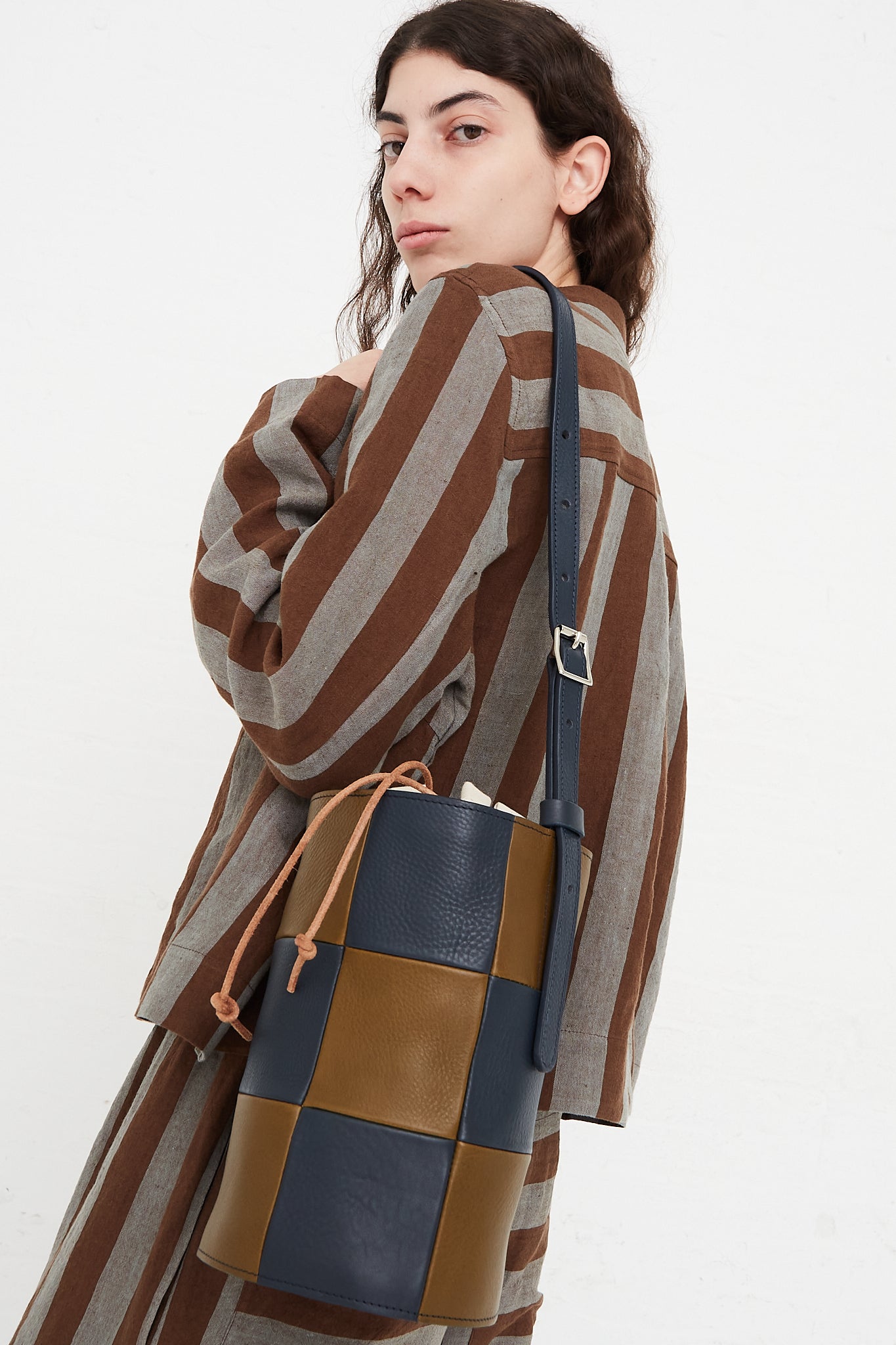 A model holding a bucket bag in a grainy patchwork leather. Features an adjustable strap with silver hardware, and an interior canvas bag with leather tie closure. Side view and upclose. Draped over models shoulder. Designed by Cawley - Oroboro Store