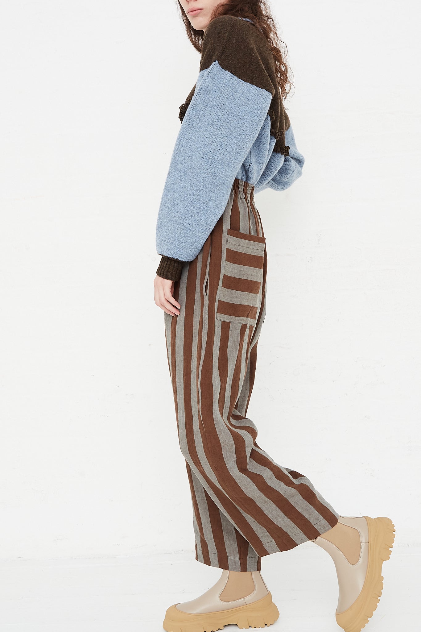 A model wearing a high waist trouser in a striped Irish linen. Brown and blue stripe. Features an elasticated waist and a wide straight leg. Full length and side view of model highlight full pant, sweater and boots. Showing pocket details. Designed by Cawley - Oroboro Store
