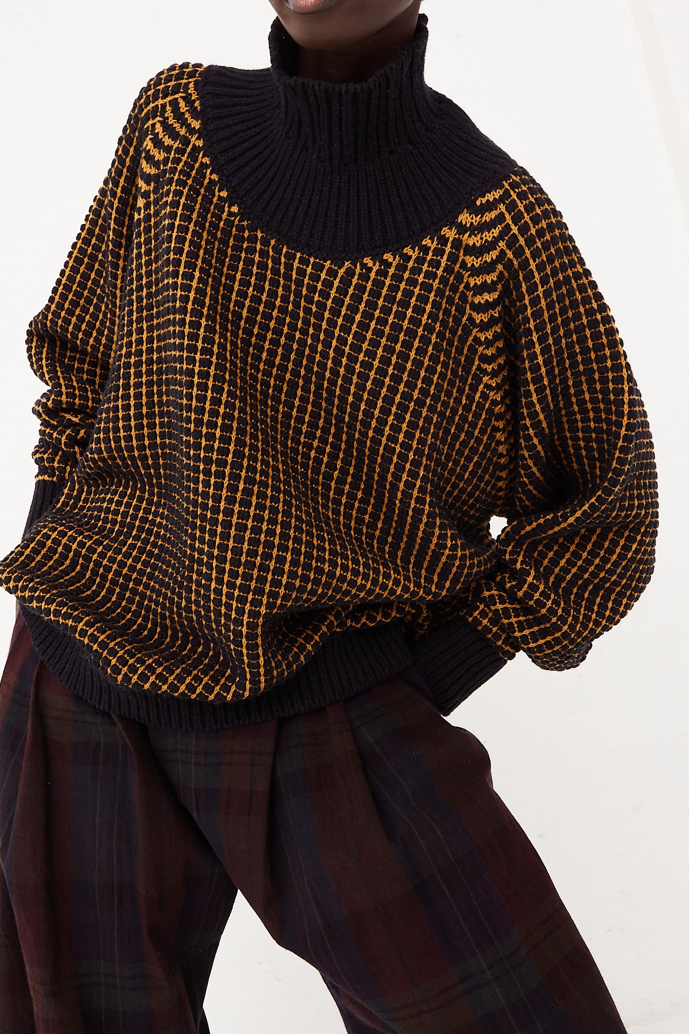 A woman is posing for a photo wearing the Jan-Jan Van Essche Turtleneck Sweater in Pitch Black Gold Lalin and plaid pants, showcasing the stylish knit design of her outfit.