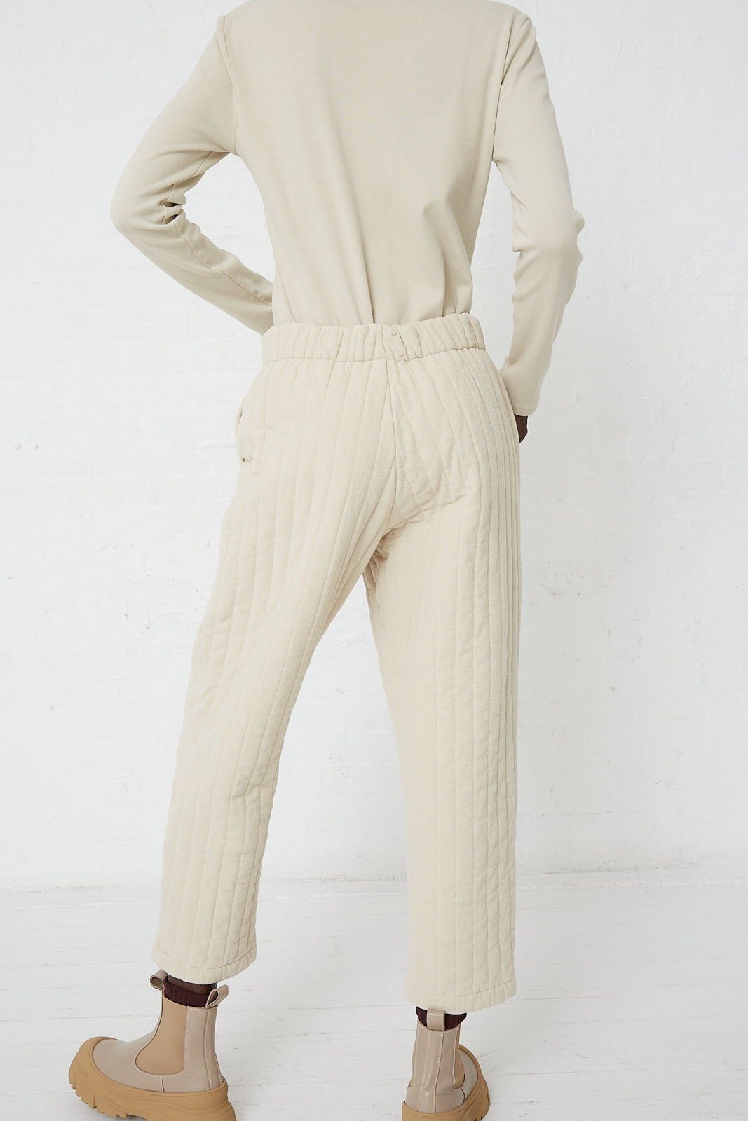 The back view of a woman wearing Black Crane Quilted Easy Pants in Ivory and a turtleneck.