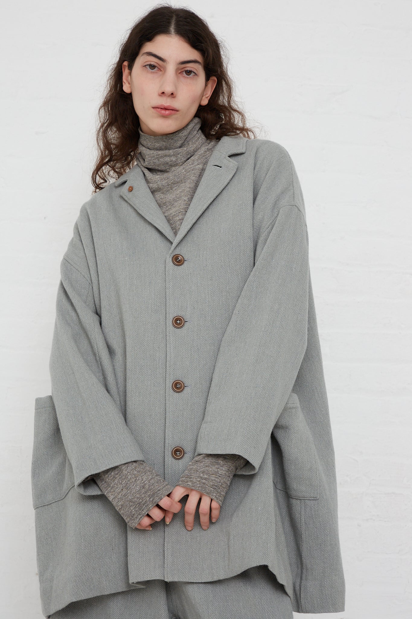 The model is wearing an oversized grey coat with patch pockets and a turtleneck made of Merino Wool Orihimedaki Jacket in Blue from Ichi Antiquités.