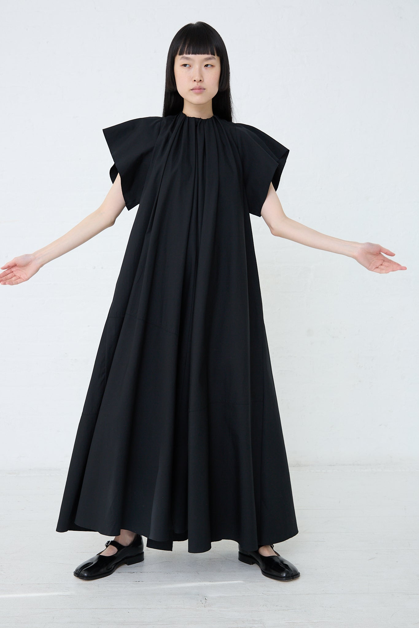 A woman in a oversized black cotton poplin MM6 maxi dress with arms outstretched. Front view and full length.