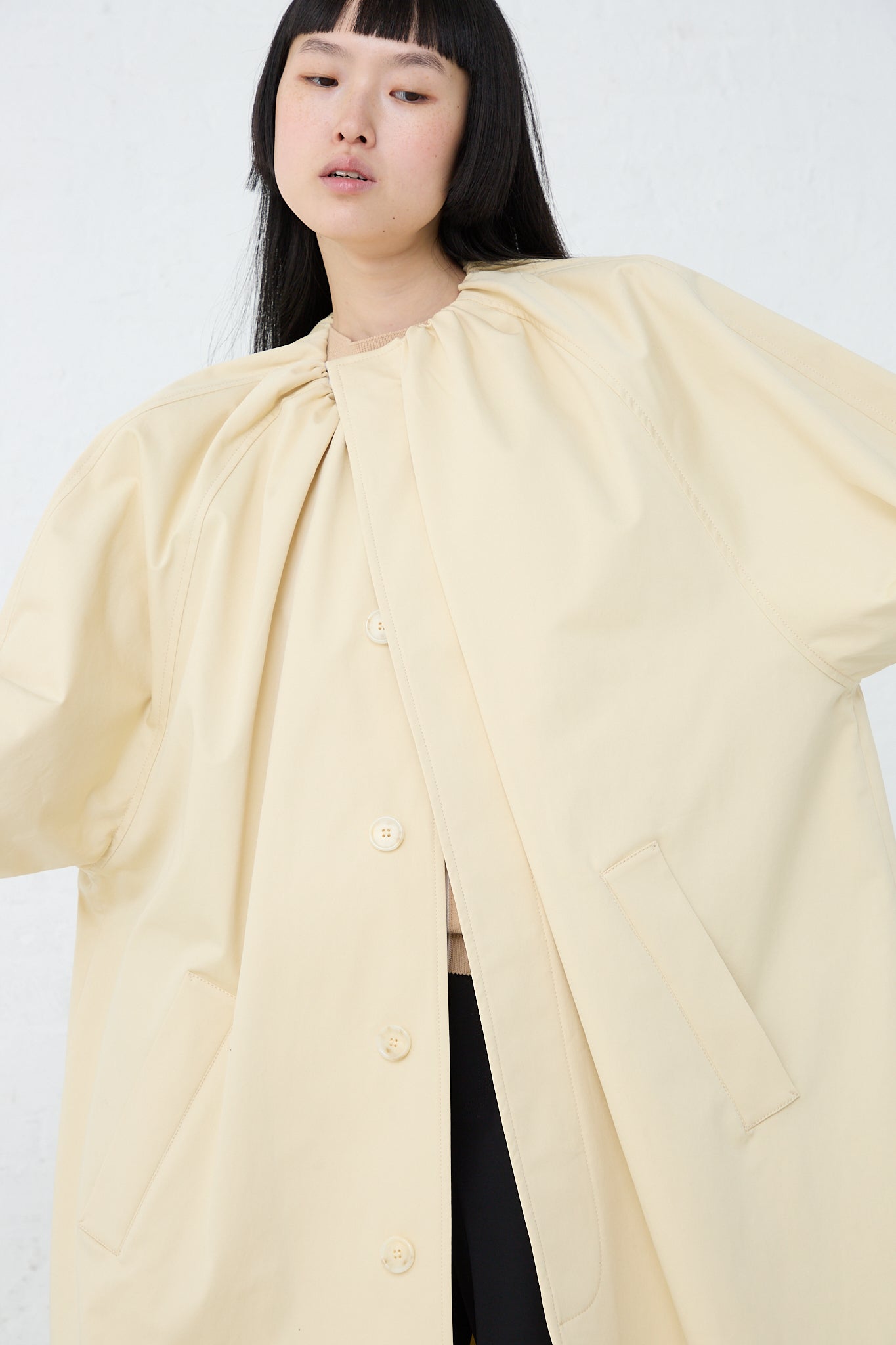 The model is wearing a MM6 collarless trench coat in pale yellow. Front view and coat is closed.
