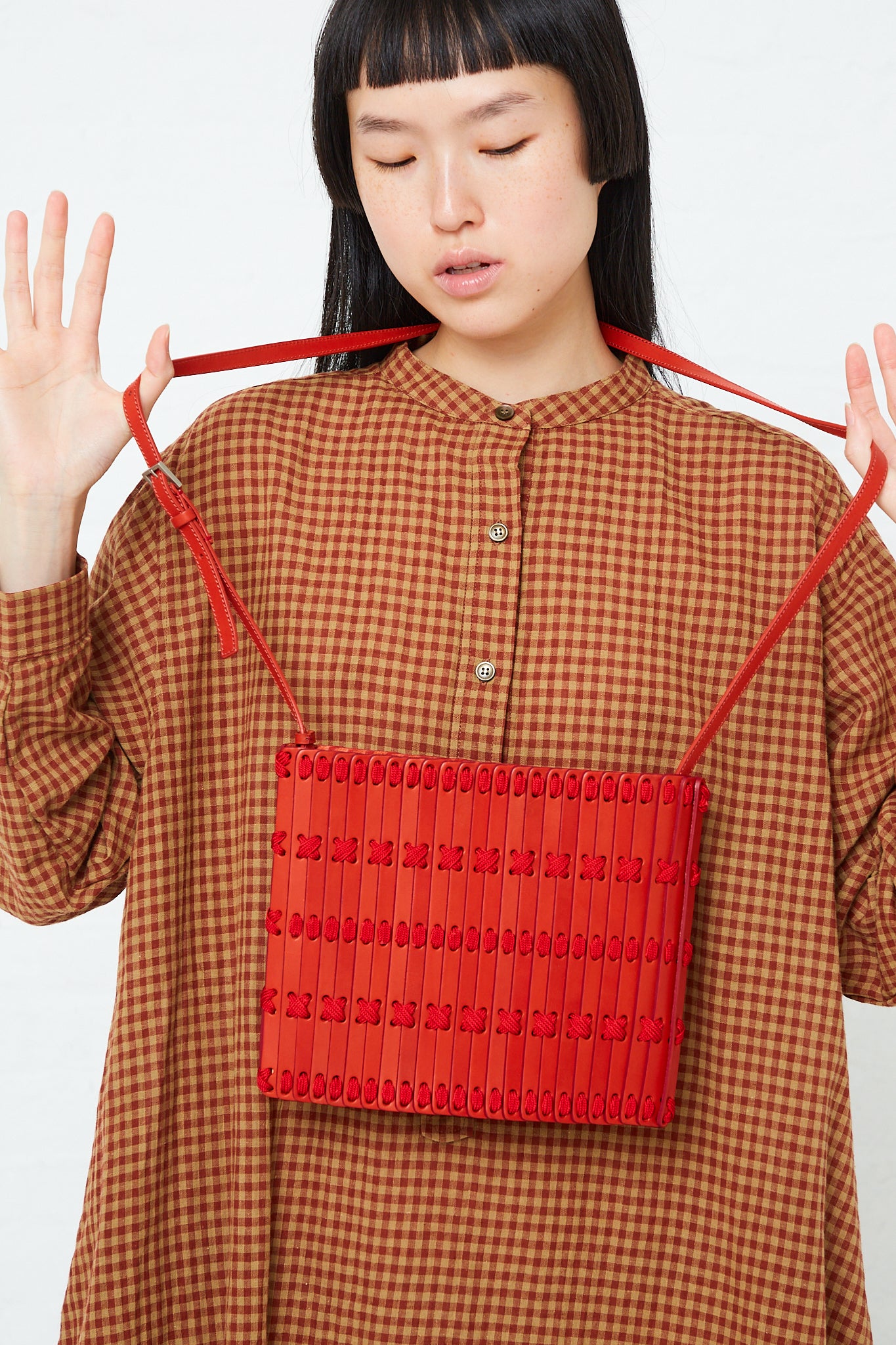 A woman wearing a plaid shirt holding a Hatori Crossbody Bag 46 in Amaranto and Ruby.