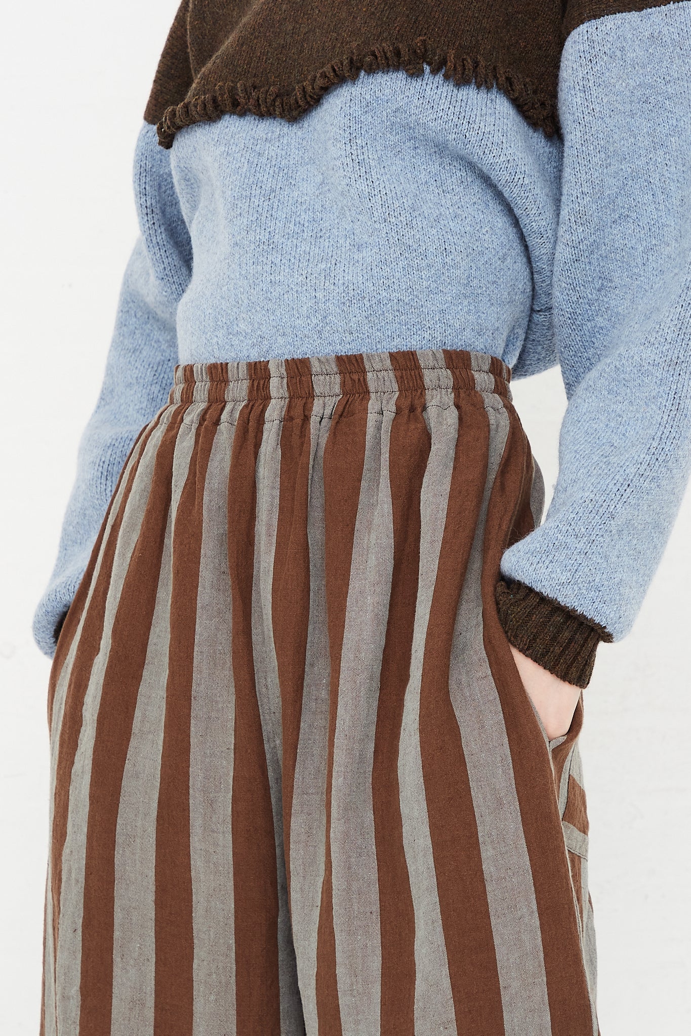 A model wearing a high waist trouser in a striped Irish linen. Brown and blue stripe. Features an elasticated waist and a wide straight leg. Front and upclose view of model highlighting pant details and sweater. Hands in pockets. Designed by Cawley - Oroboro Store