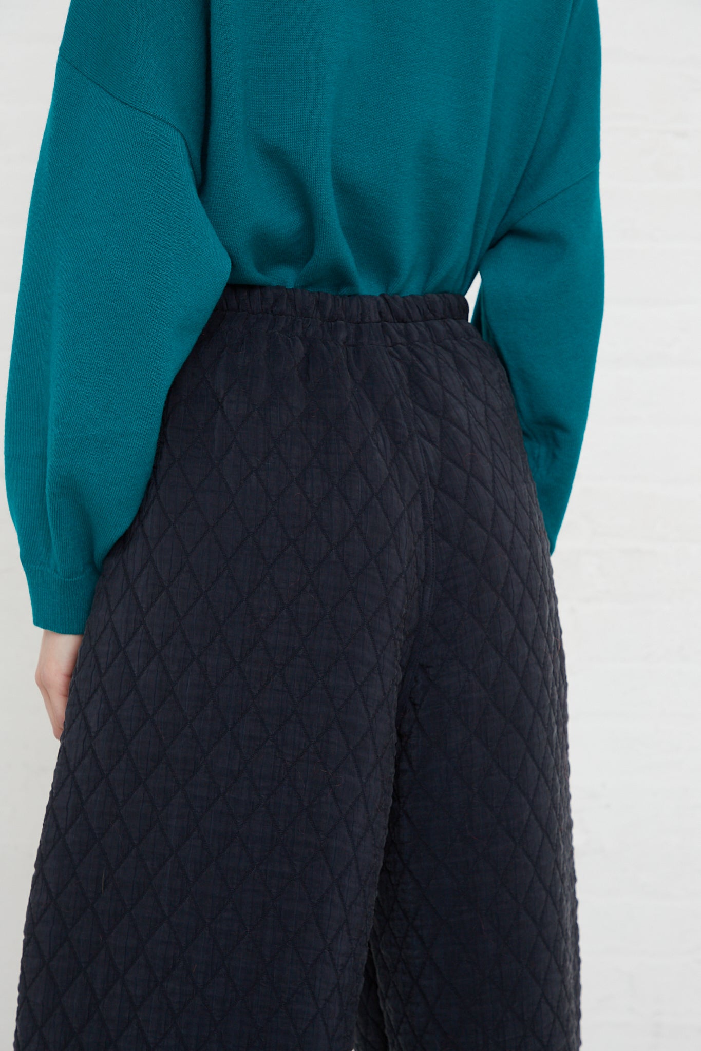 The back view of a woman wearing a teal sweater and Cordera's Quilted Curved Pant in Black with a high rise waist.