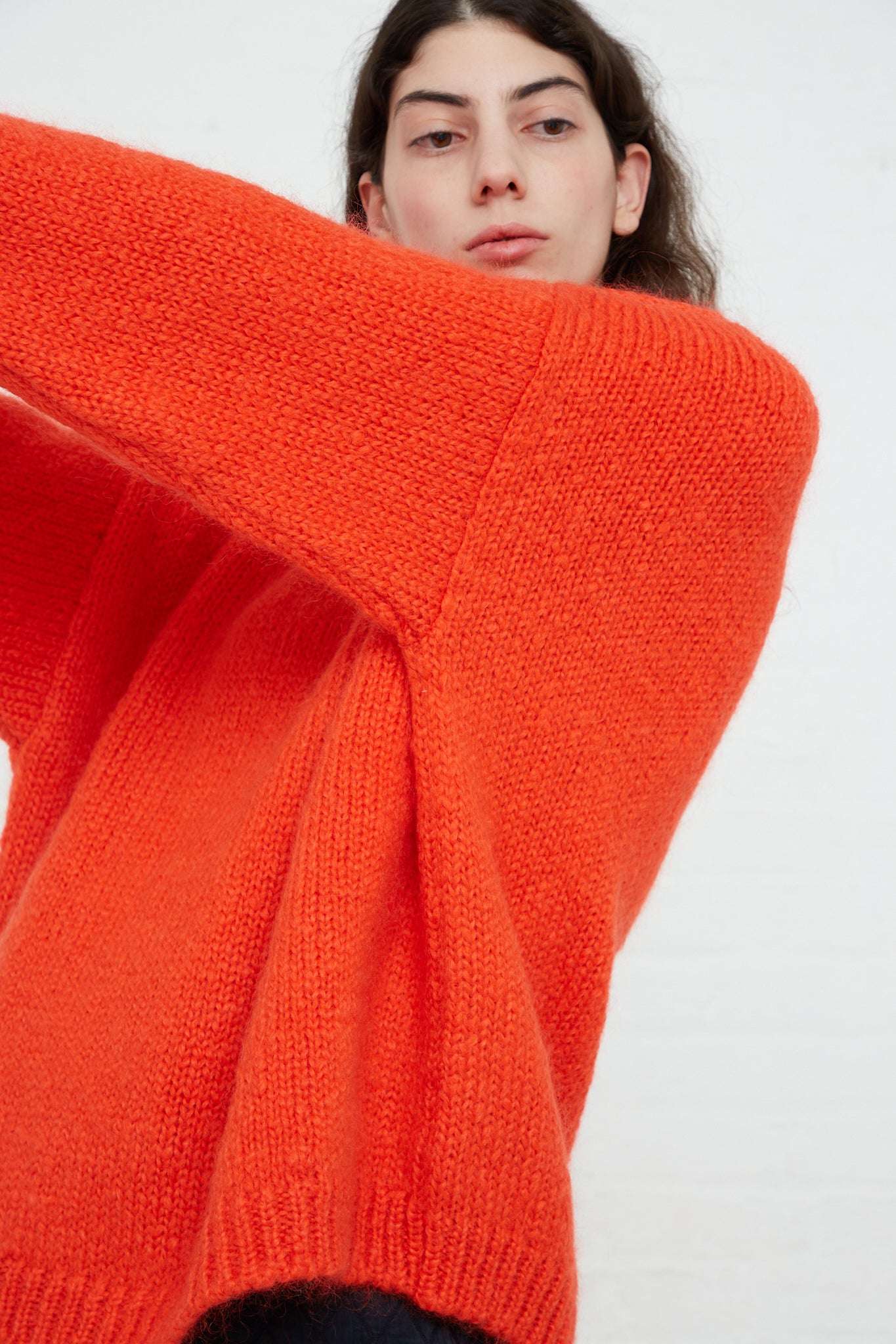 A woman in an oversized Cordera Mohair Sweater in Tangerine, made from a mohair blend, is posing in front of a white wall.
