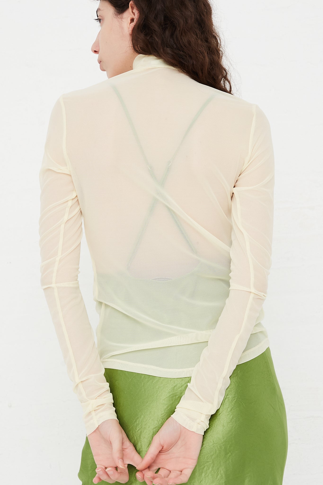 The back view of a model wearing a green skirt, designed by Nomia's Long Sleeve Mesh Mockneck in Ivory.