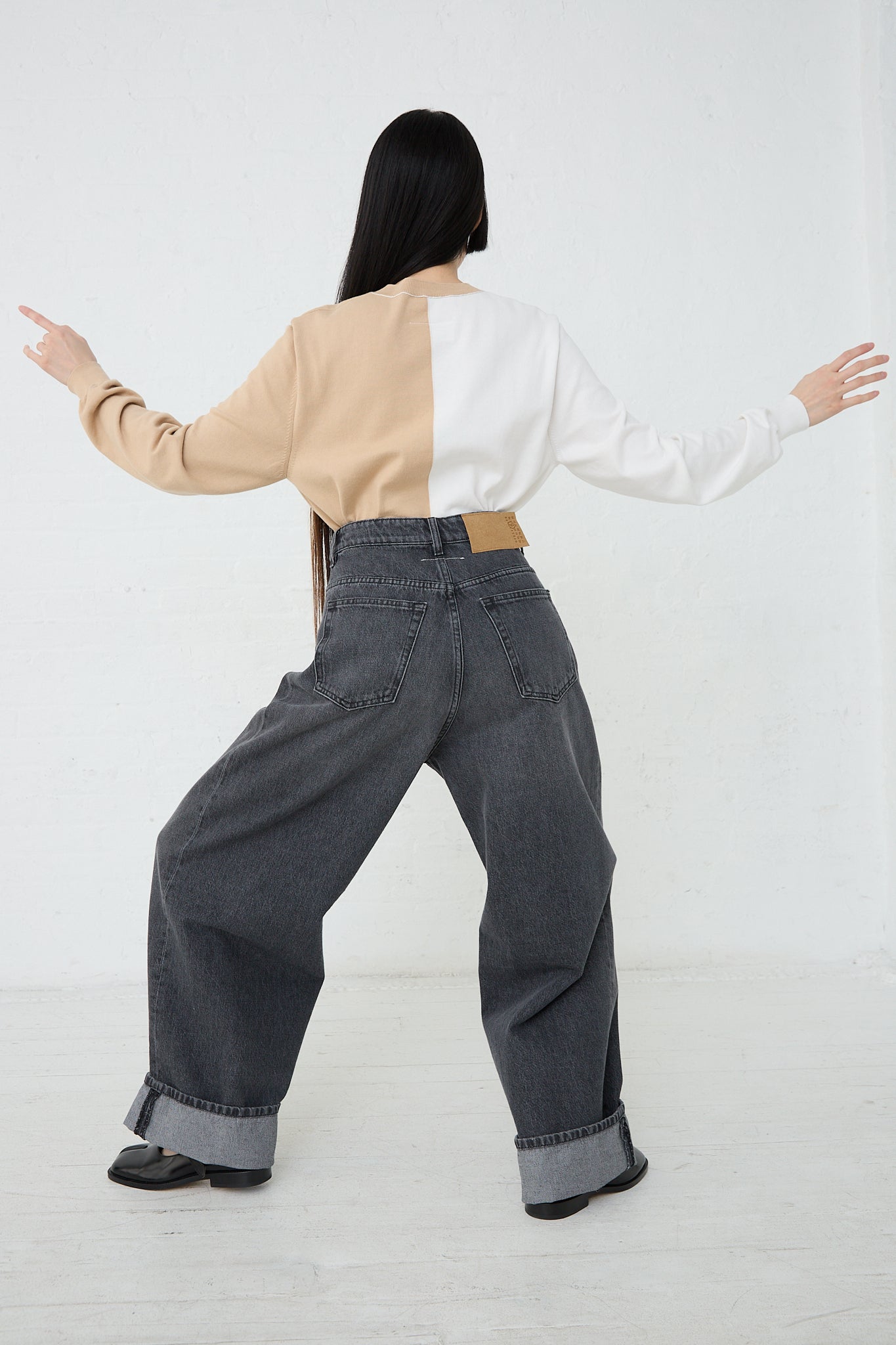 A woman is standing in a white room with her arms outstretched, wearing MM6's 5 Pocket Pant in Grey denim. Back view and full length.