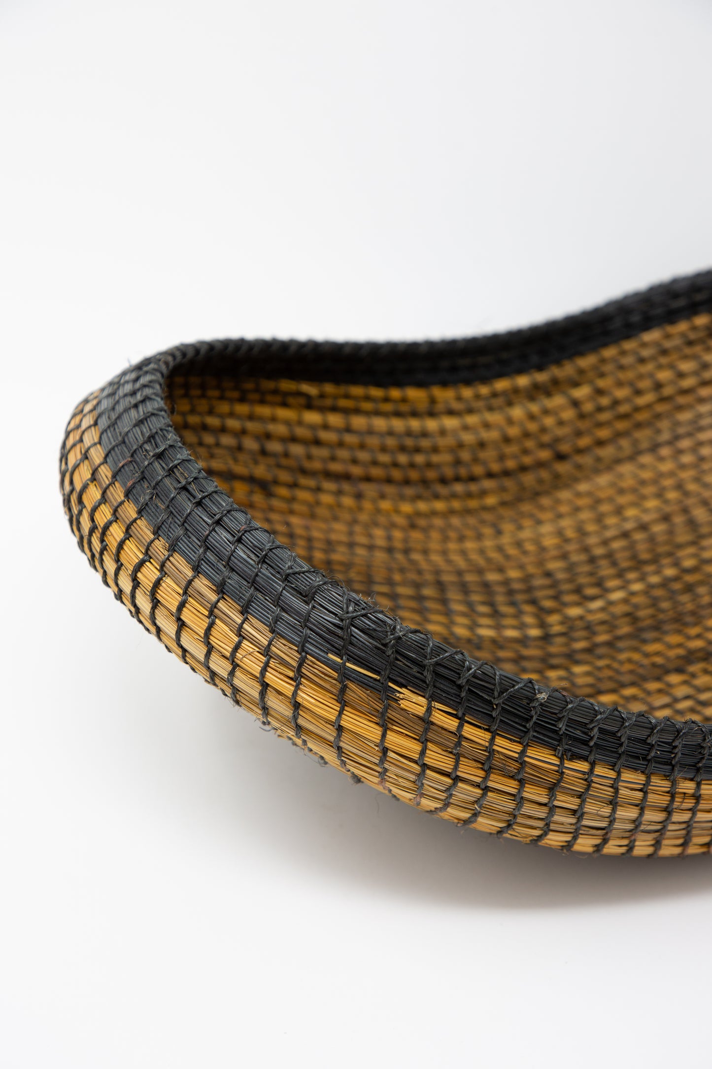 A traditional Plaza Bolivar Asopafit Canoe Basket, woven in black and brown, on a white surface. Up close view. 