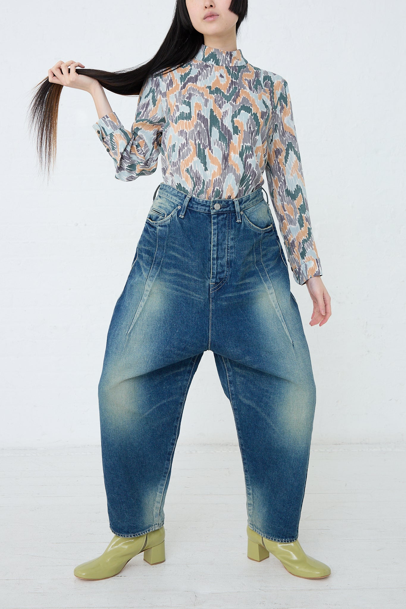 A woman in a floral blouse and denim pants with relaxed fit, wearing the Mina Perhonen Always New Tapered Pant in Blue. Front view. Model is holding her hair in her right hand.