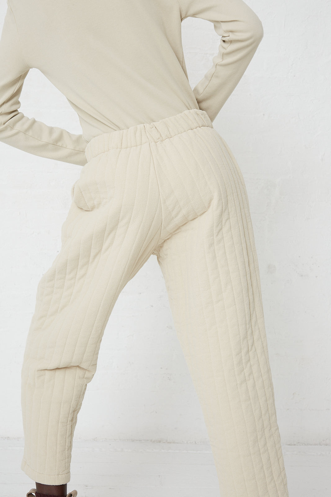 A model is standing in a white room wearing a turtleneck and Black Crane's Quilted Easy Pants.