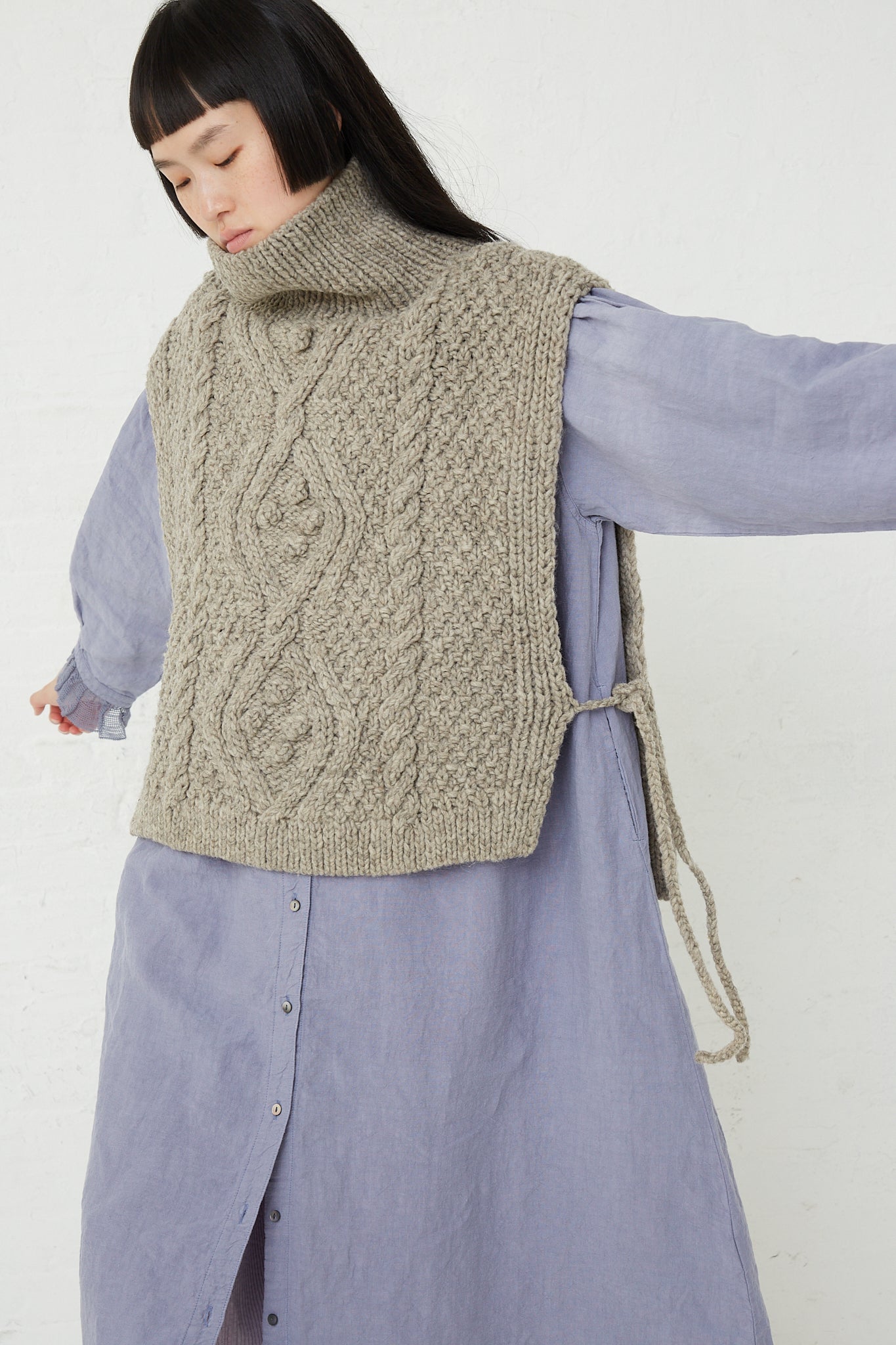 A woman in a blue dress is wearing the "Peruvian Wool Hand Knitted Cable Stitch Vest in Beige" by nest Robe, an open-side turtleneck vest knitted with ethically raised sheep wool.